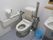 フタが無いタイプのトイレには、ベージュ色の背もたれと手すりが設置されている。右下に、掃除用のブラシとゴミ箱が置いてある。