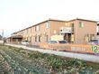 地上２階建ての施設は、茶色の外壁でシックな雰囲気である。建物の左側には駐車スペースがあり、その横には広い畑がある。