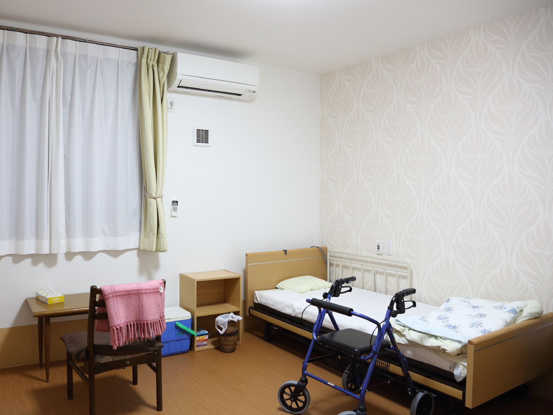 布団が敷かれたベッドの前に、青い歩行補助器が置かれている部屋