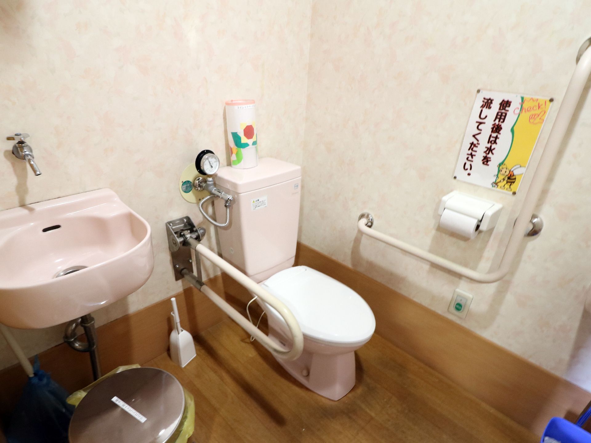 右奥の便座の両側に手すりが敷設されており壁際にはナースコールも見える。左側は手洗い場になっておりゴミ箱もある。