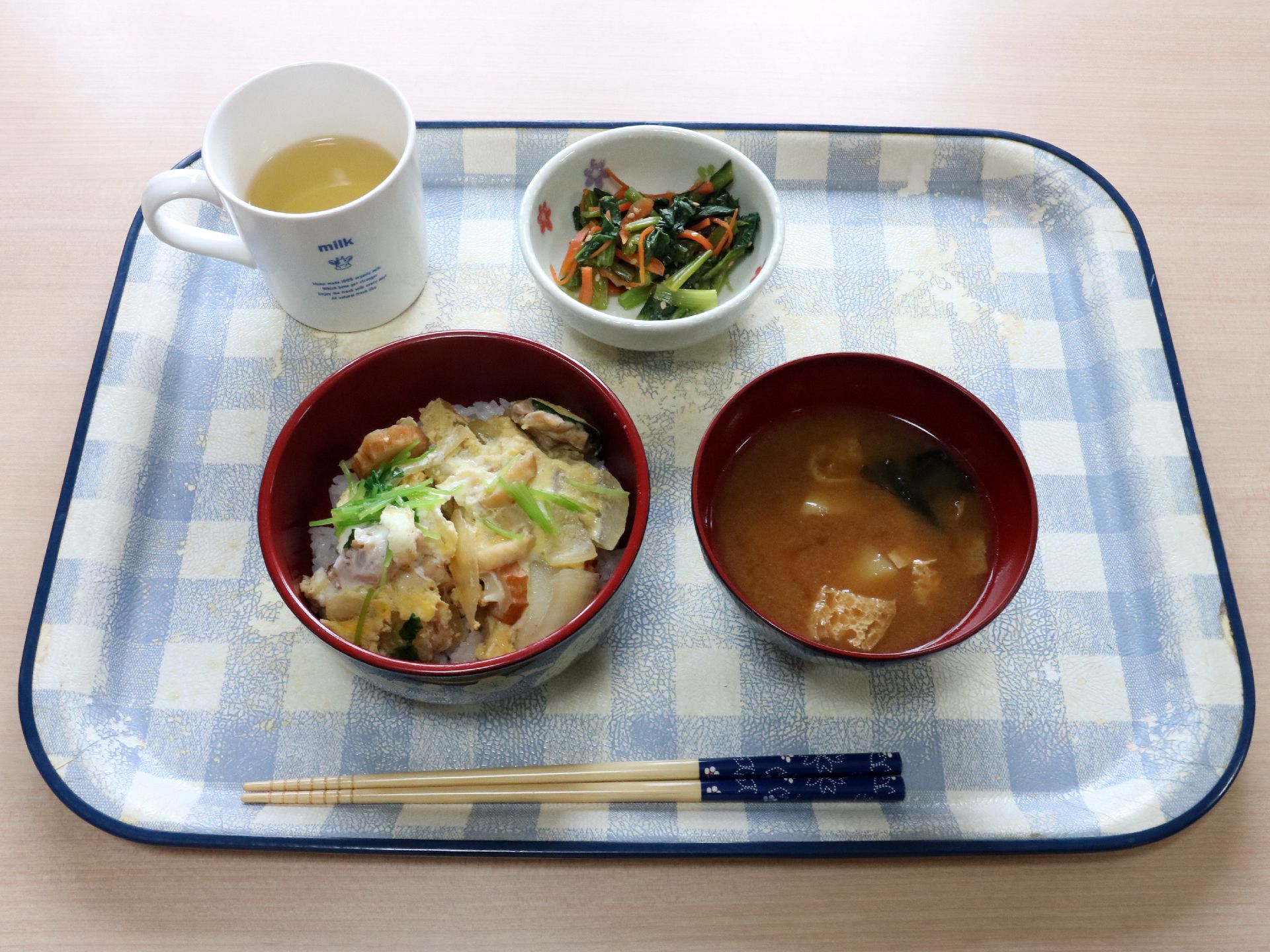 チェック柄のトレイに健康を考えた食事が乗せられている。卵とじの丼ものと小鉢、味噌汁にお茶が提供されている。