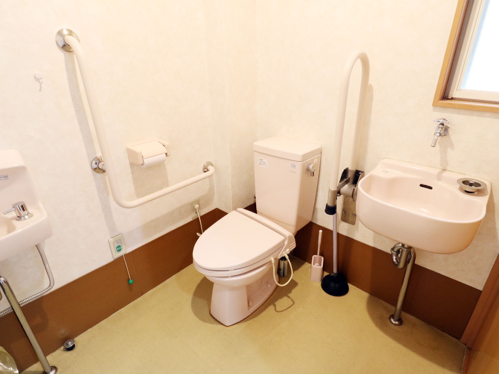 左側の壁にはL字型の手すりがついておりその下には呼び出しベルもみえる。便座をはさんで反対側には可動式の手すりもついている。