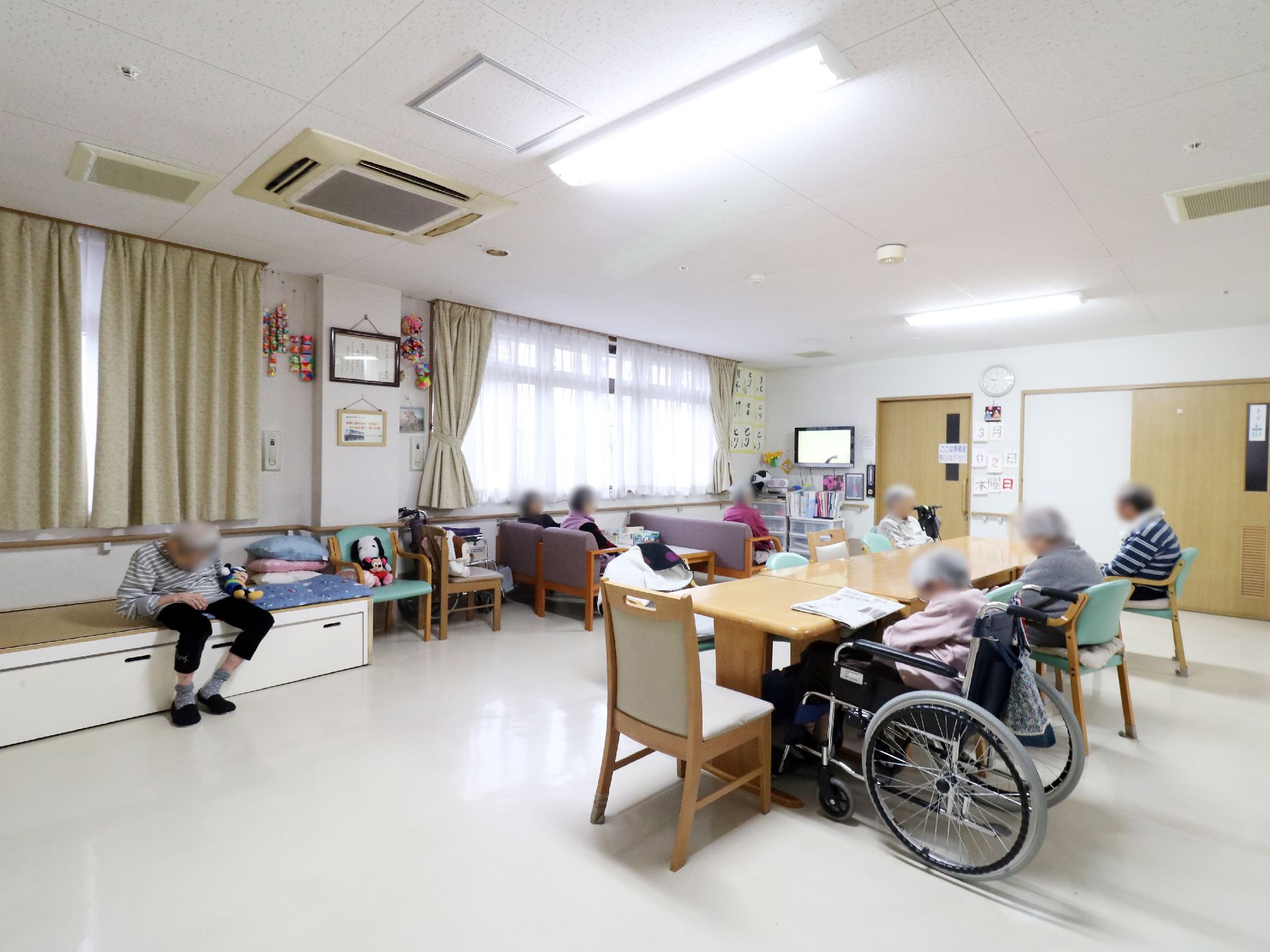 広々とした部屋では木製の長いテーブルに椅子や車椅子の入居者が座っている。他にも窓際のテレビをみている人たちも見える。