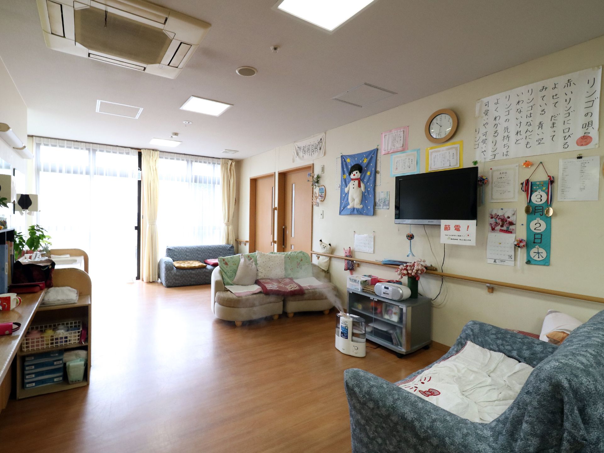 窓から陽光が差し込んでおり、天井には空調がついている。右手の壁にはテレビがありまわりにいろいろなものが貼り出されている。