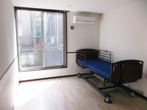居室の壁際に、高さが調節できる介護用ベッドを備えている。上部にエアコンを設置し、正面にはガラス戸があり明るい空間である。
