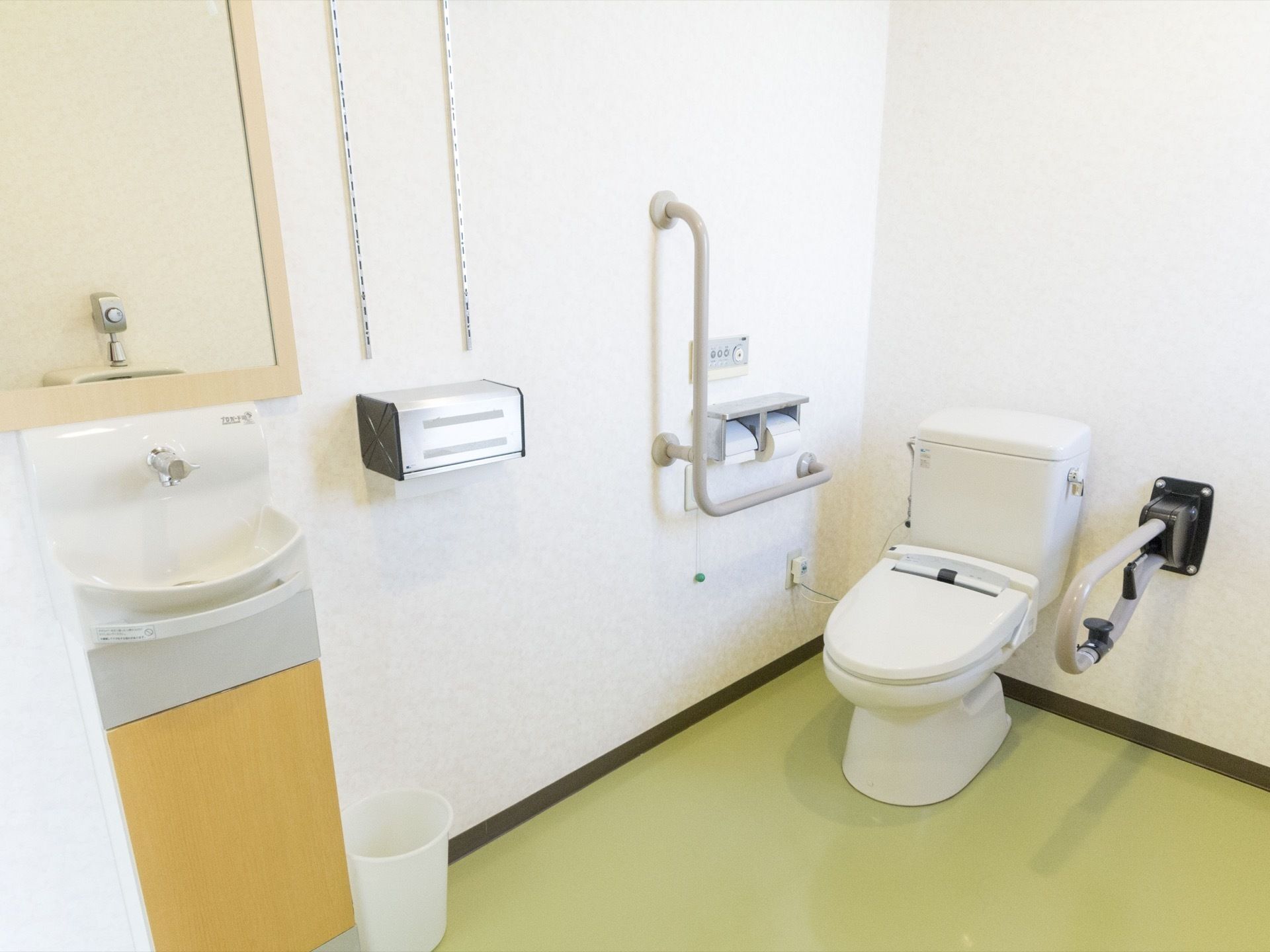 洋式トイレの個室内で、手洗い場も併設されている。便器の右壁にはＬ字型の手すりがあり、左手には可動式の手すりがある。