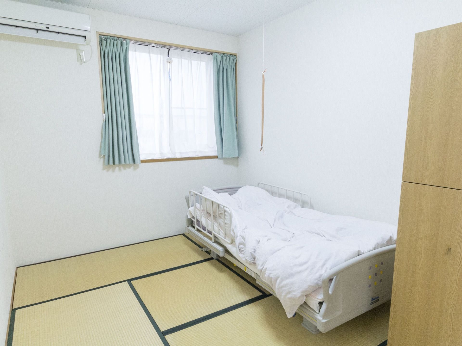 ある居室の様子。部屋の奥に窓があり、水色のカーテンが付いていて、レースカーテンが敷かれている。エアコンがあり、畳の上にベッドが置かれている。