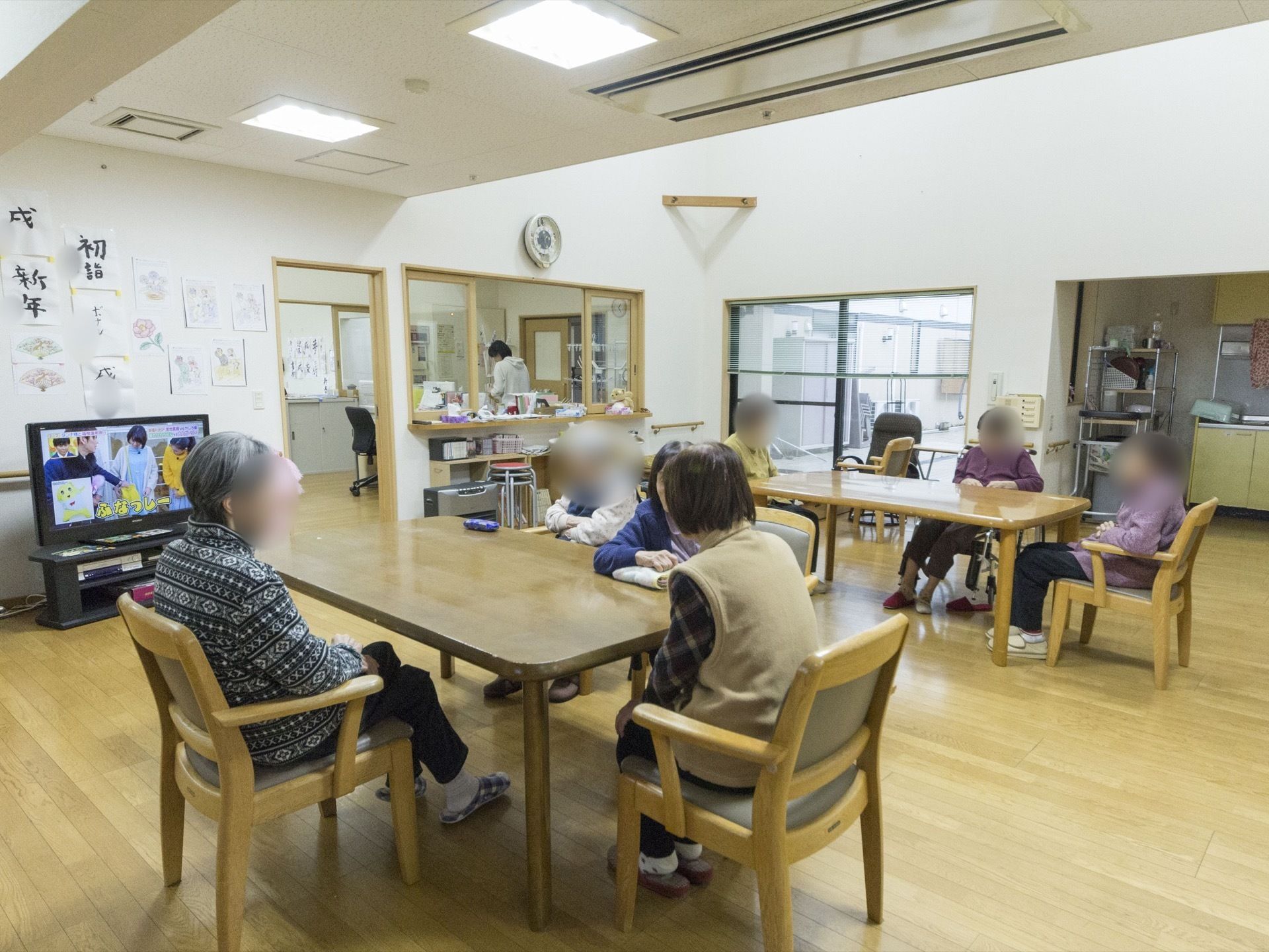 リビングの一角にテレビが設置されている。その後ろには、書道作品などが掲示されている。室内で、入居者様たちがテレビ鑑賞や会話を楽しんでいる。