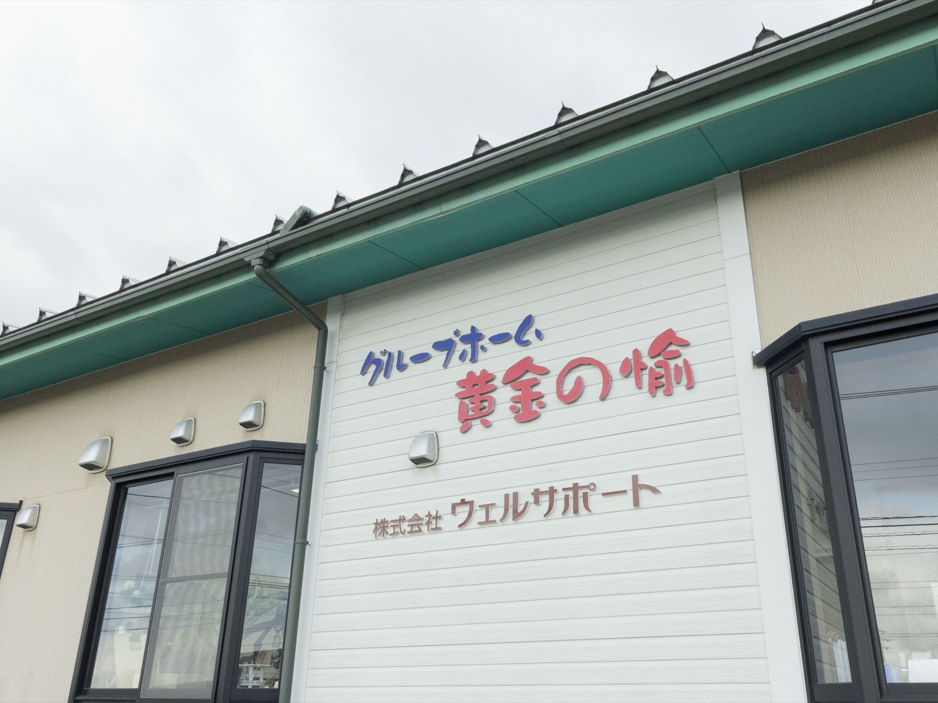 施設の建物は１階建てである。横長の建物で、施設名が表示されている両側には出窓が２つずつ並んでいる。建物の前に車が駐車している。