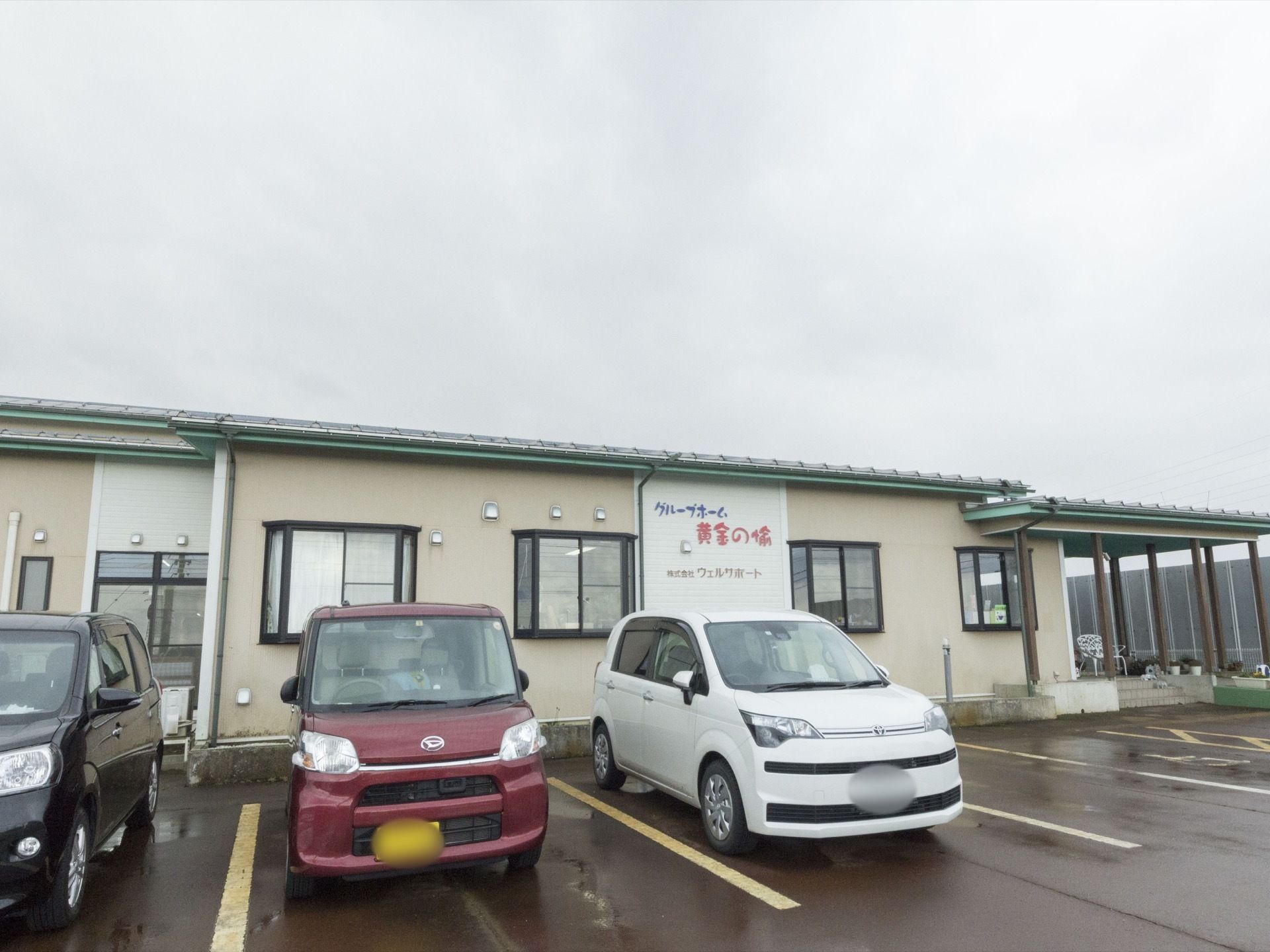 施設は横長の建物で、その外壁沿いに駐車スペースが並んでいる。建物の右端にはテラスがあり、花が飾られ椅子が置かれている。