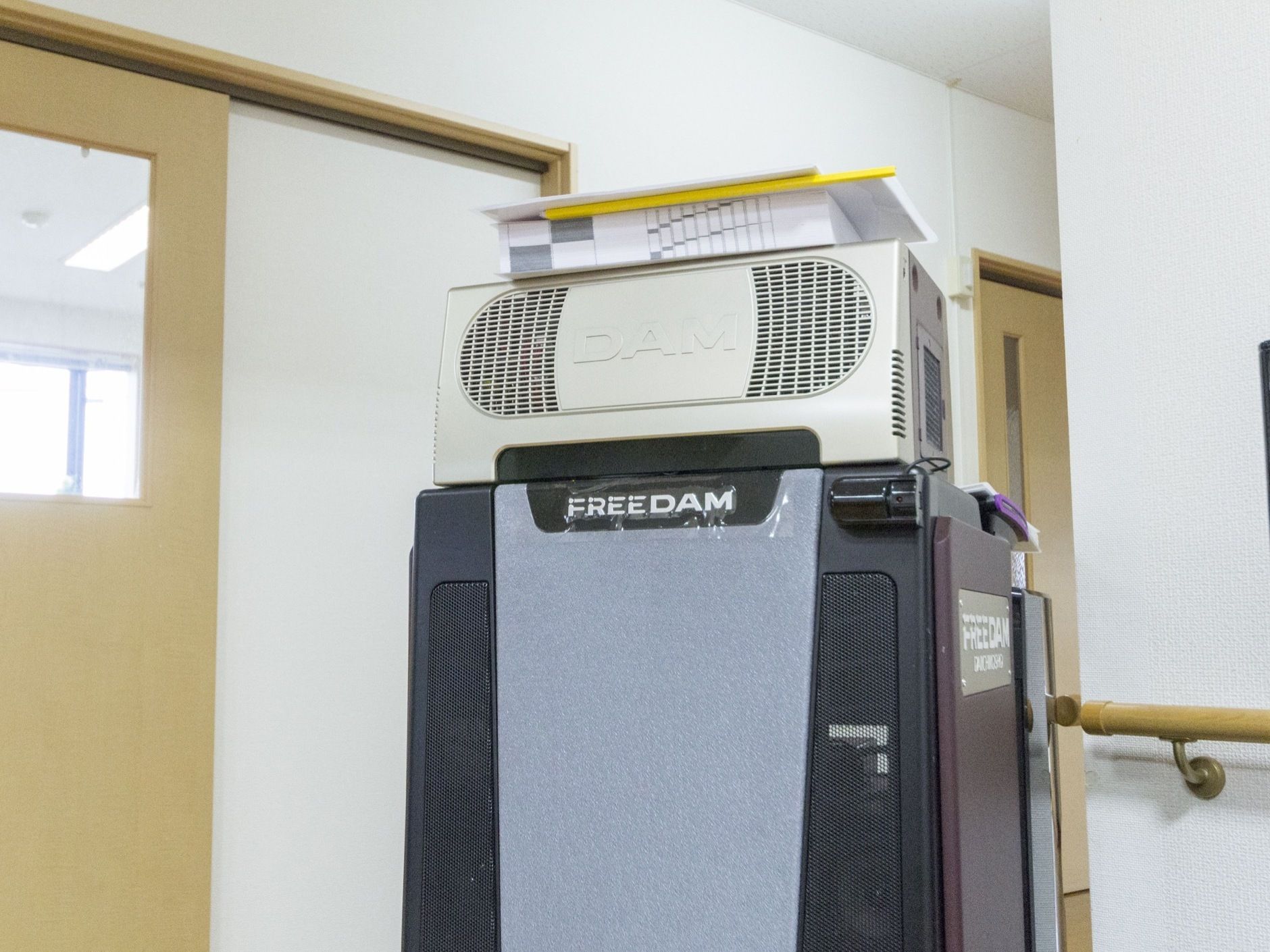施設内に完備されている、カラオケ機器。大きな機械の上に、歌本が載っている。機器は壁の近くにあり、手すりが見える。
