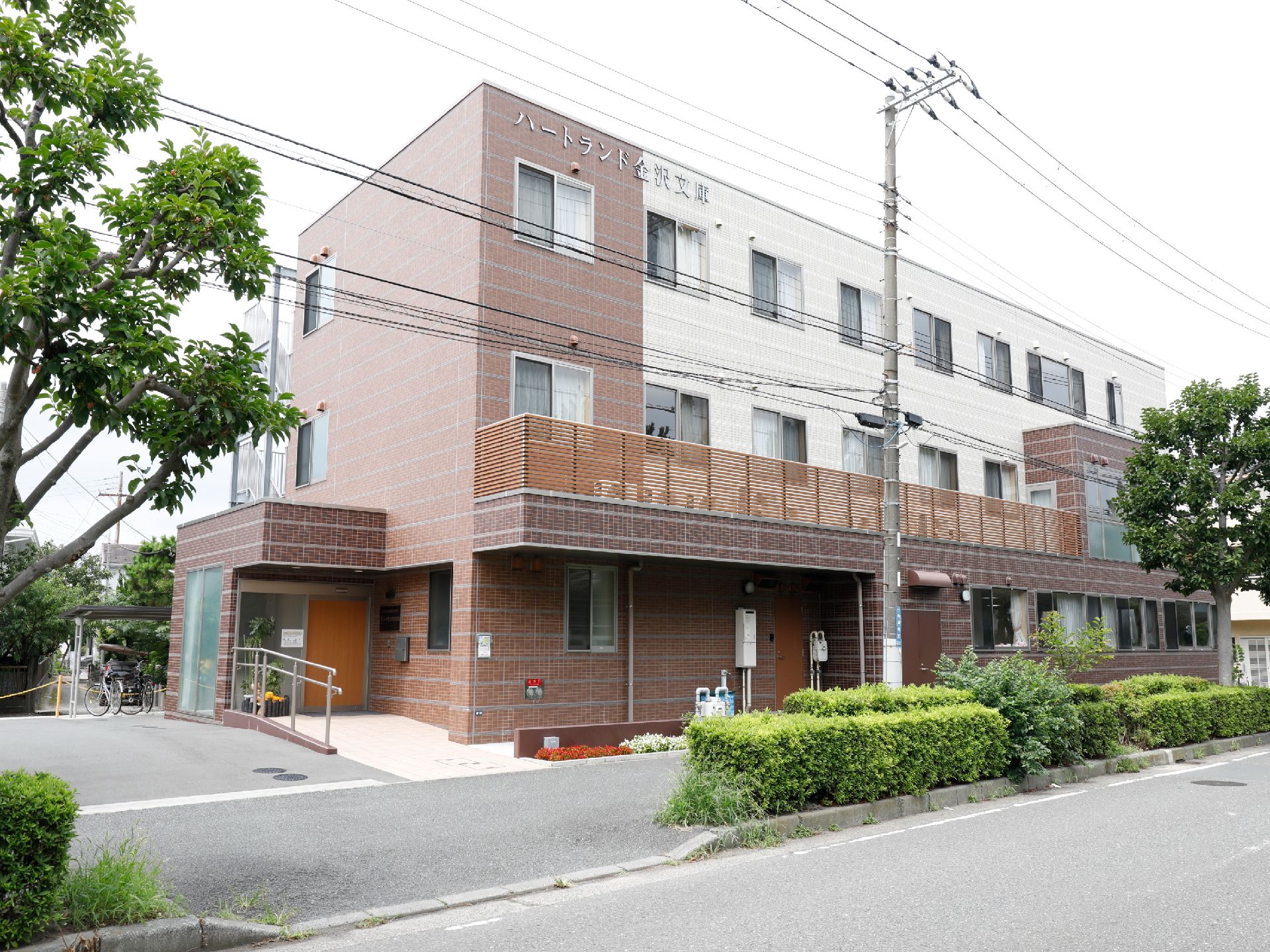 白色とダークブラウンの外観で、建物の端上に施設名が表示してある。建物は歩道に面しており、傍には植栽が施され、その向こうは大通りとなっている。