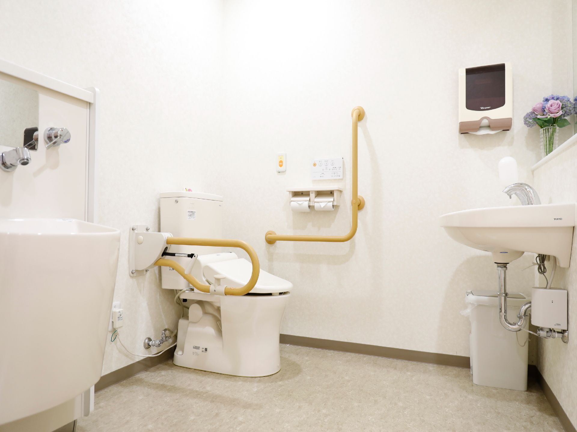 白色の壁に薄いグレイの模様入りの床のトイレで、便器の傍には手すりが設置されている。正面には洗面台が設置してある。