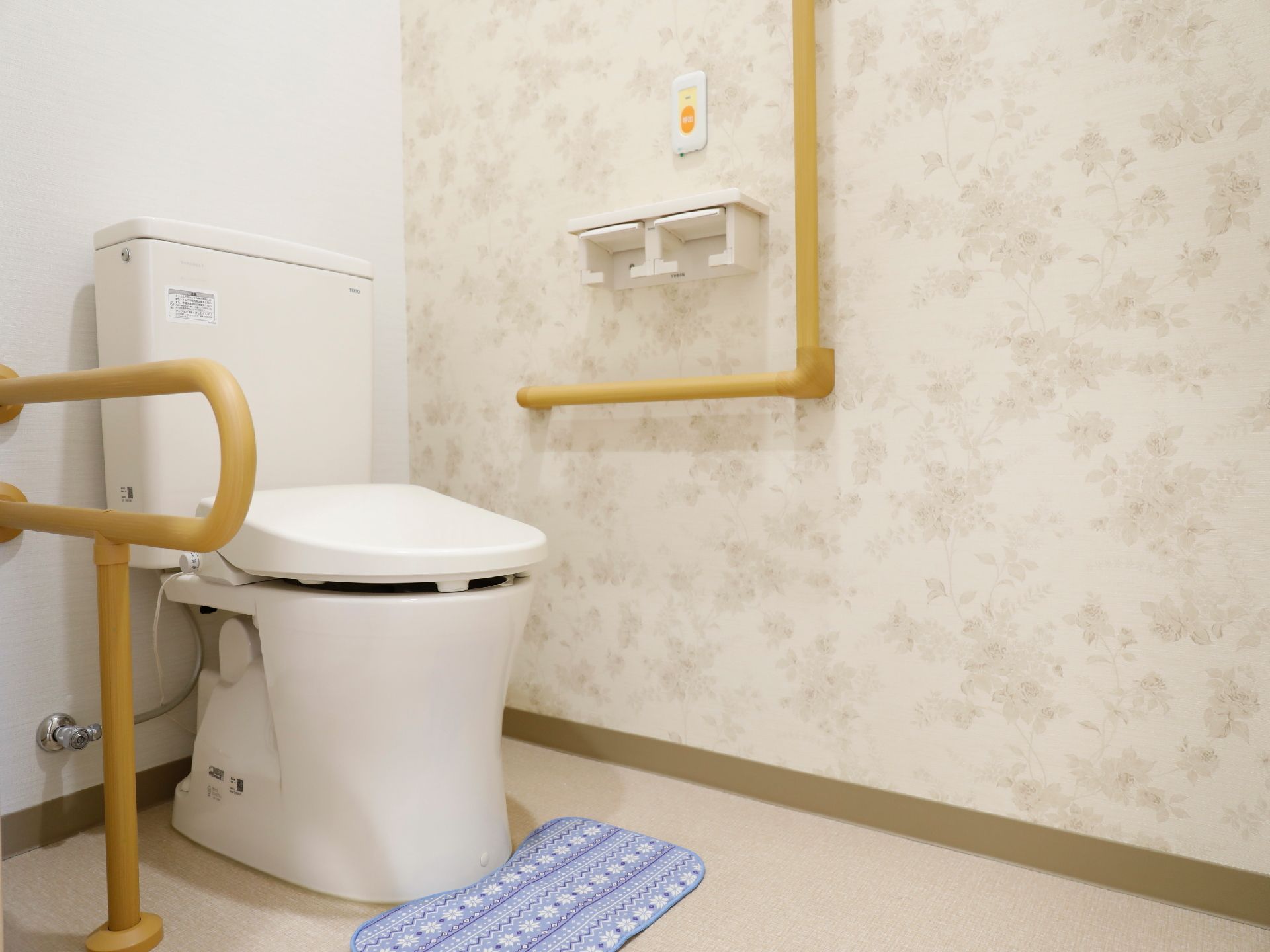 白色と花模様の壁にベージュ色の床で、便器の両側には手すりが設置されている。また、横壁にはナースコールが取り付けてある。