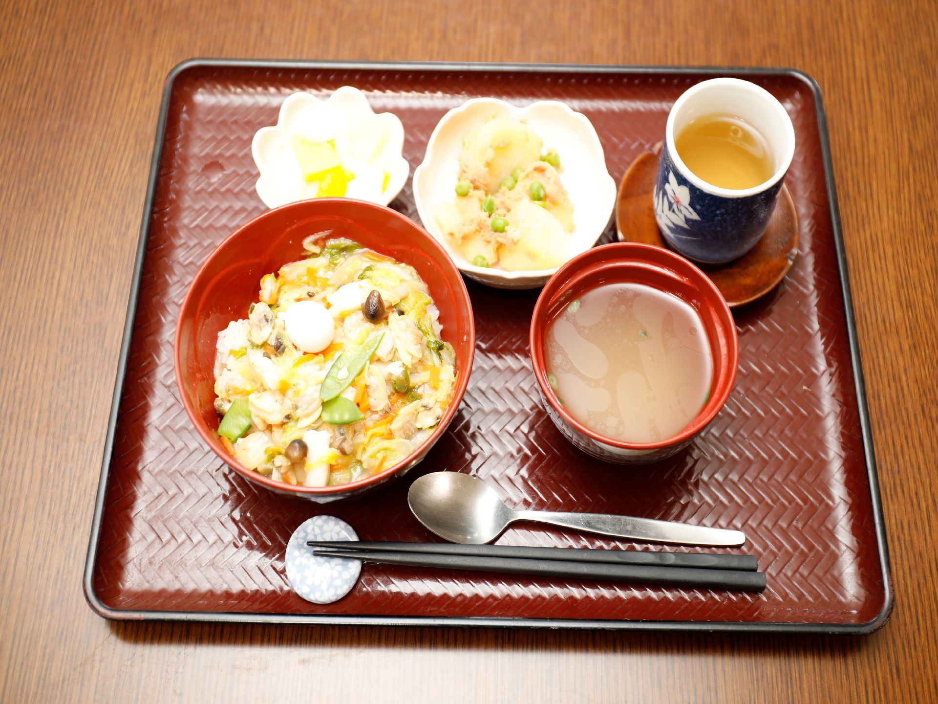 茶色のトレイの上に丼物と汁物、小鉢が2つにお茶が添えられている。手前に箸とスプーンが置いてある。1食の献立である。