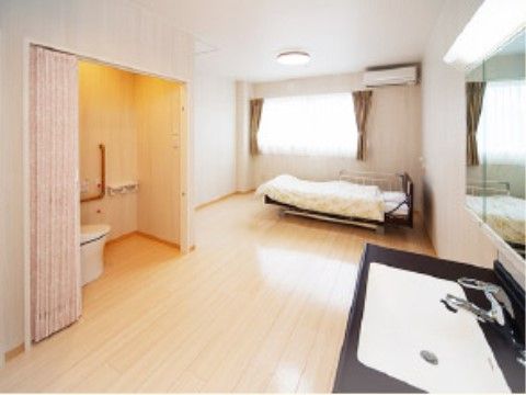 居室には窓がありトイレや洗面台が完備されている。介護用ベッドが用意されている。壁の色は白色基調で床はフローリングになっている。