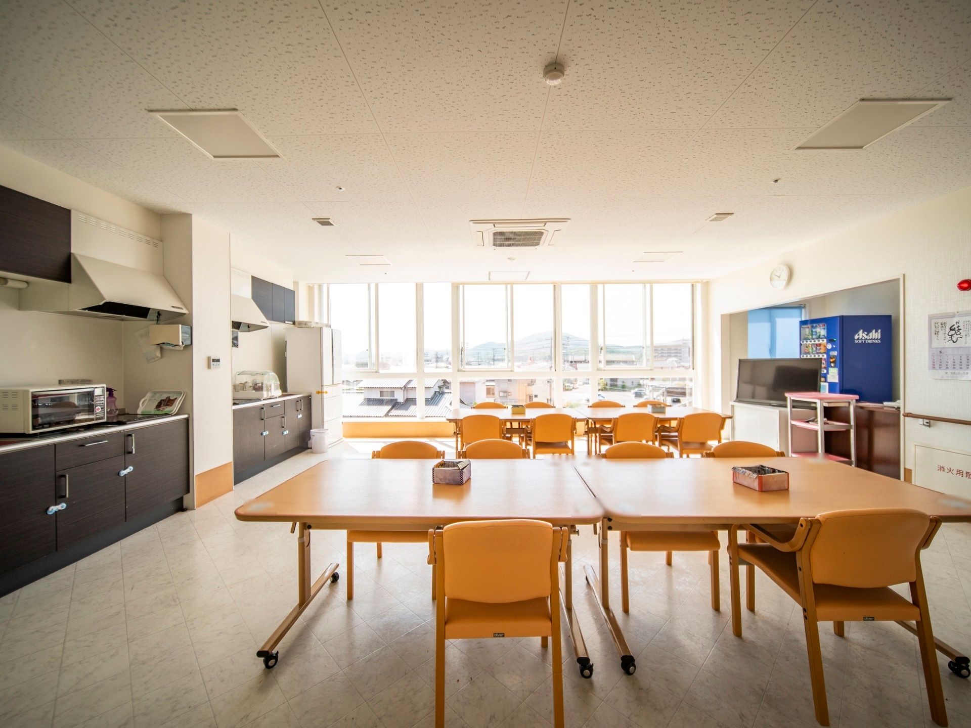 食堂にテーブルが４つと、椅子が多く置かれている。窓に向かって左側にキッチンがあり、レンジフードが２か所についている。