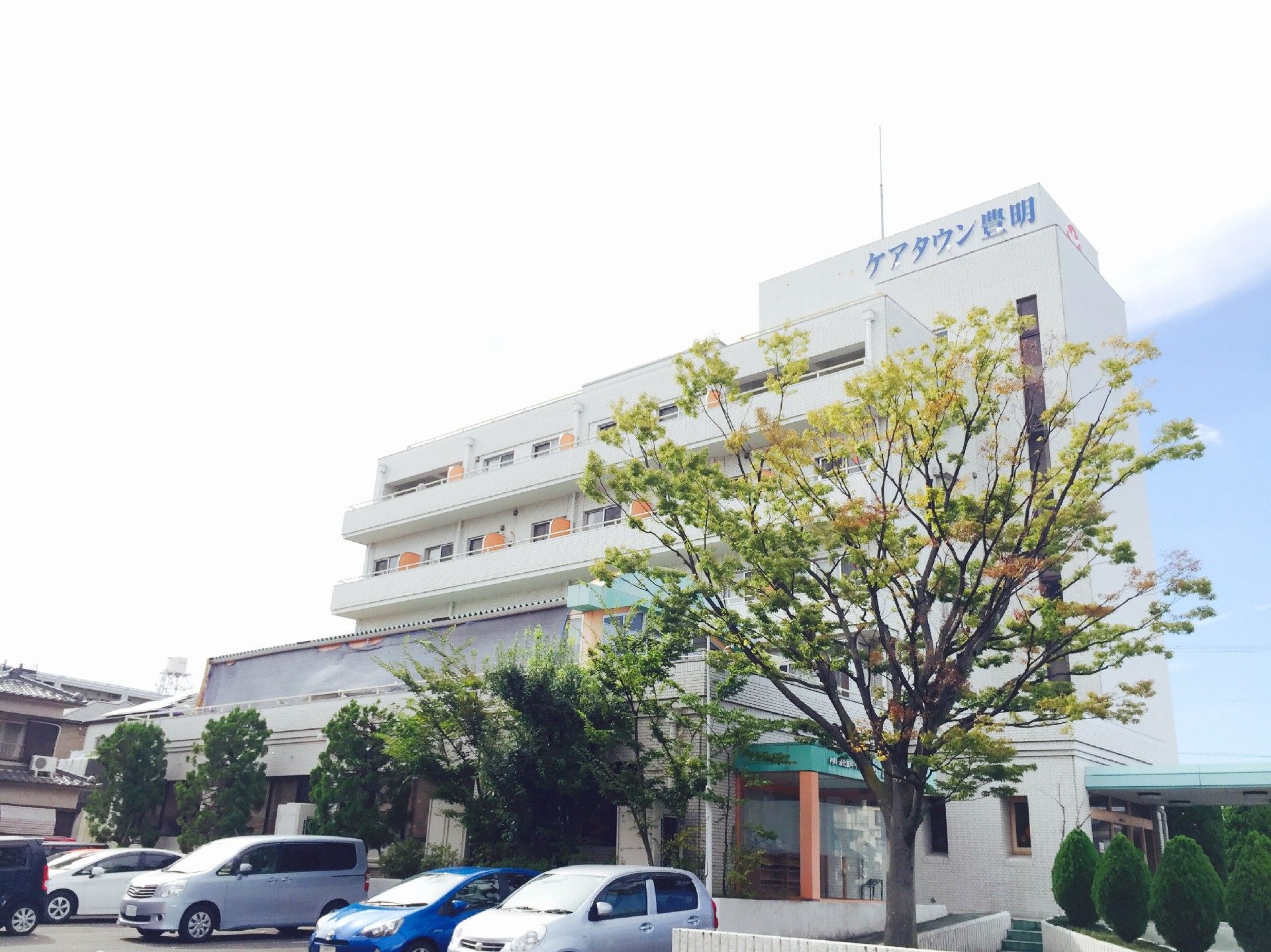 一階のロビー前に背の高い木が、数本ある。木の前には駐車場が用意されている。エントランスの屋根は緑、柱はオレンジである。