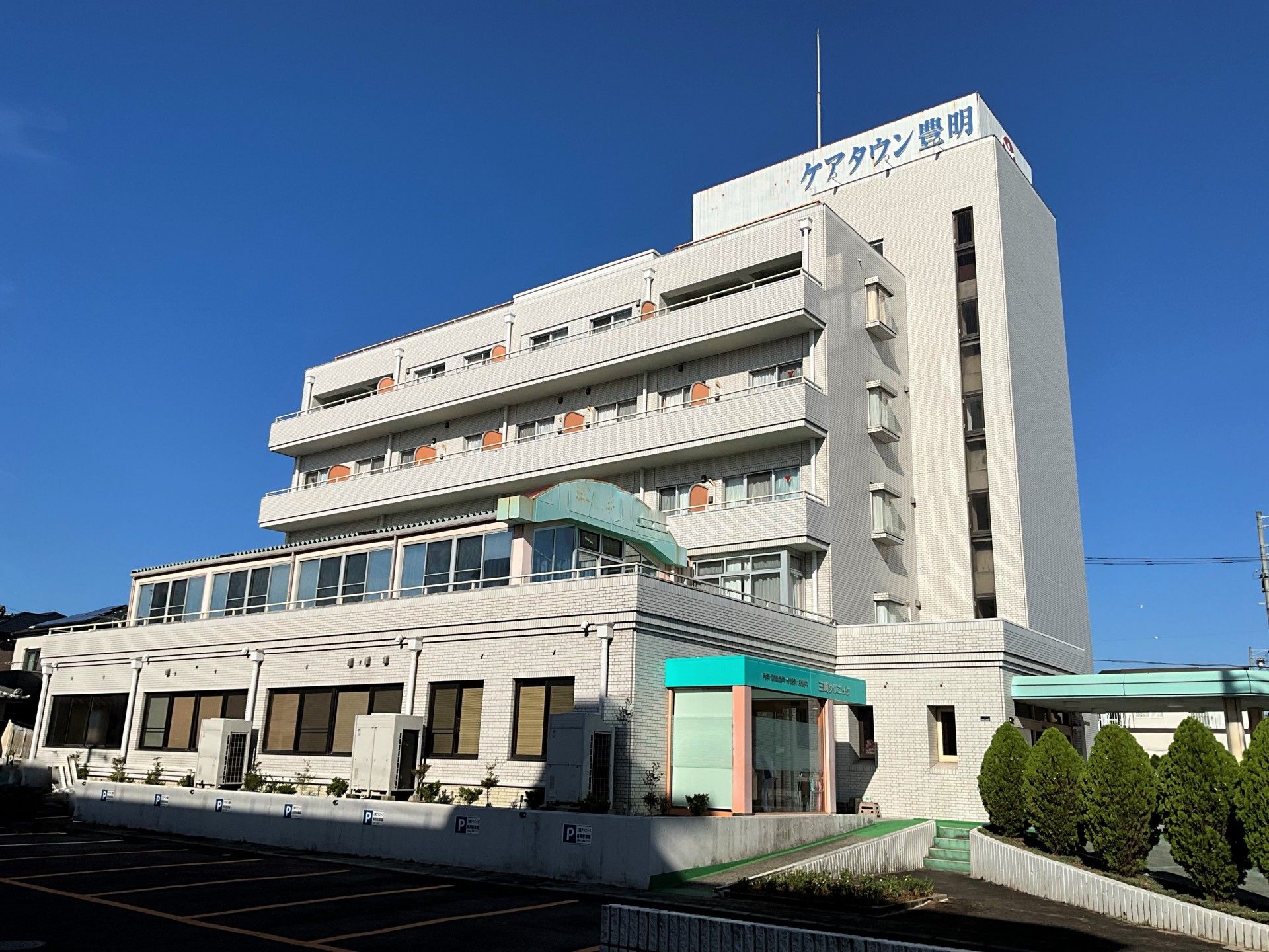 一階のロビー前に背の高い木が、数本ある。木の前には駐車場が用意されている。エントランスの屋根は緑、柱はオレンジである。