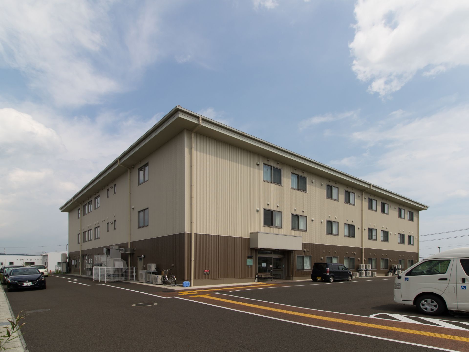 住宅型有料老人ホームの建物は、ブラウンを基調とした外観になっている。また広々とした敷地内には駐車スペースが確保されている。