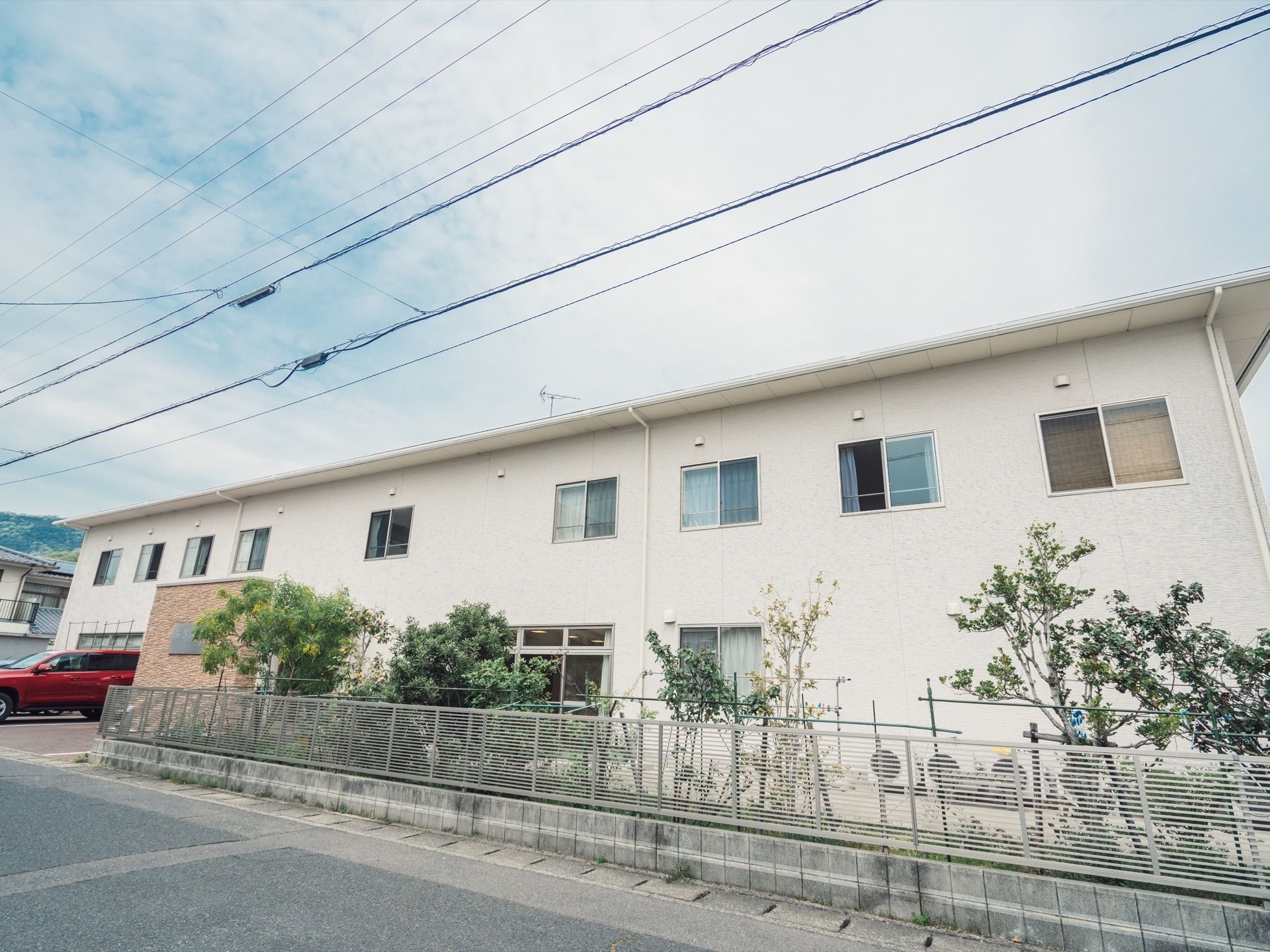 二階建ての白い外壁の建物が写っている。建物の周りはフェンスで囲われており、フェンスの内側には植物が植えられている。