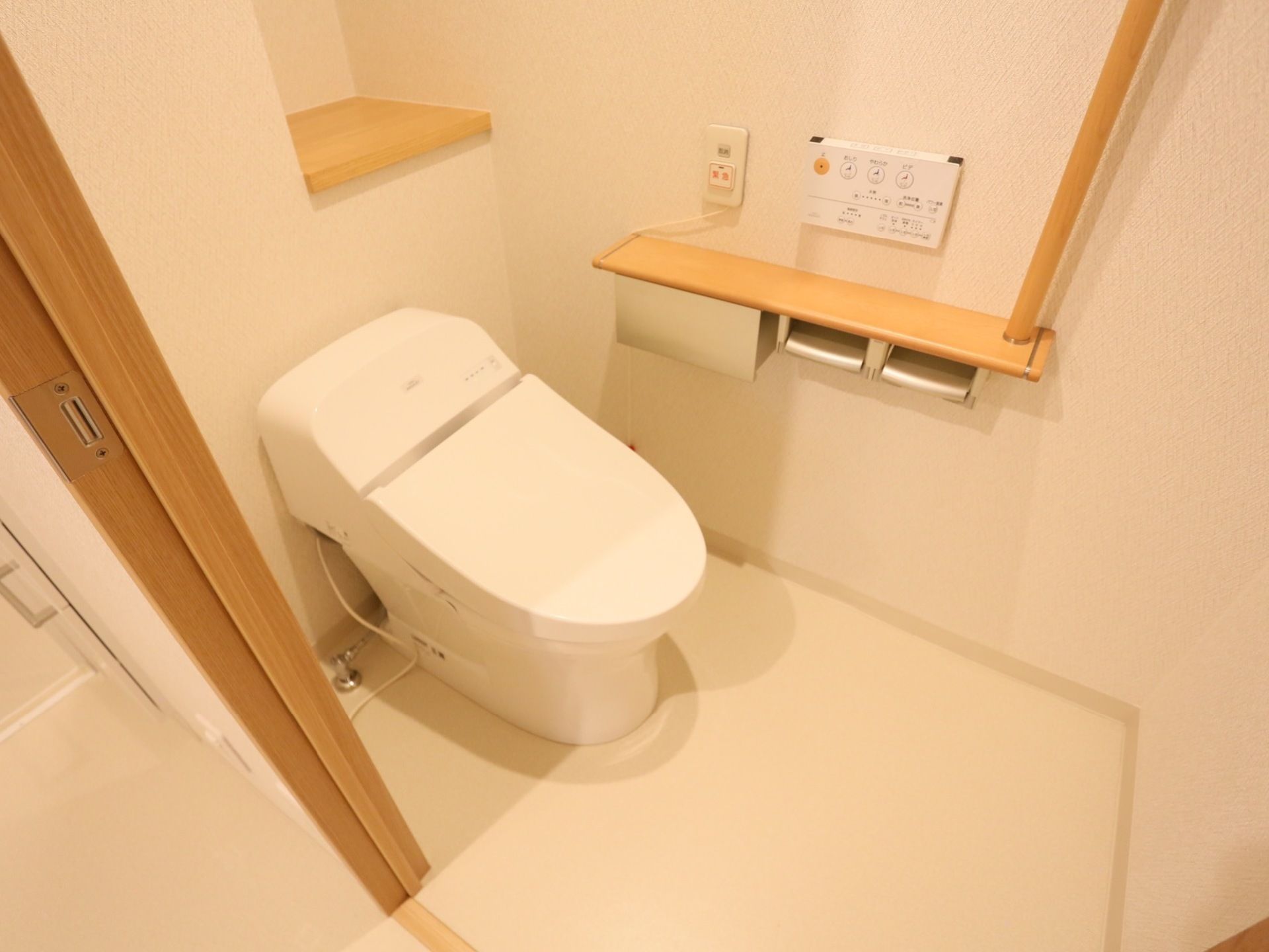トイレには、シャワー洗浄機能付きの新型便座が設置されている。壁面には、非常用コールボタンと便座操作パネル、手すりが並んで取り付けられている。