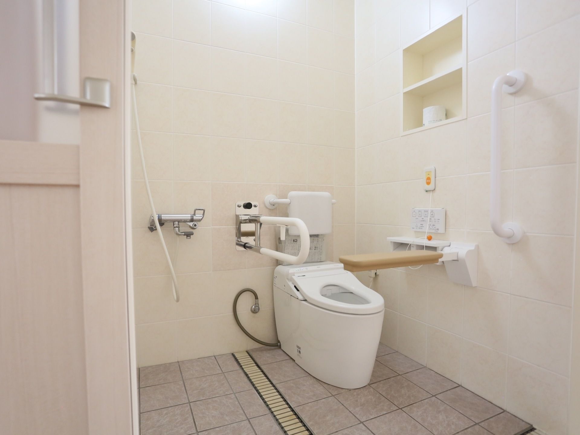 背もたれと手すりがついた洋式トイレで、横にはシャワーが設置されている。上の棚にはトイレットペーパーが用意されている。