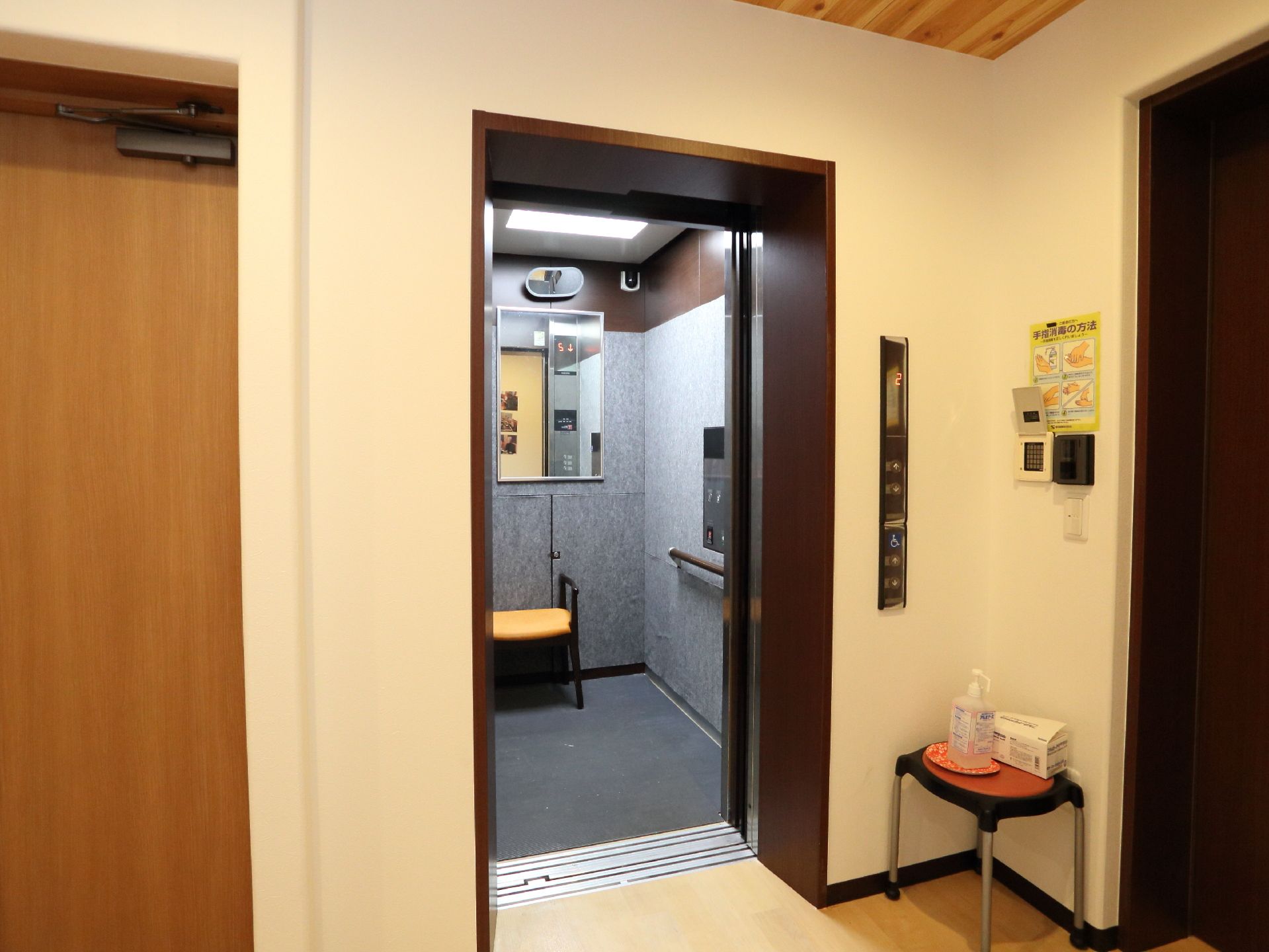 施設のエレベーターには手すりが備わっている。また、利用者が楽に利用できるように椅子が置いてある。出入り口前には消毒液がある。