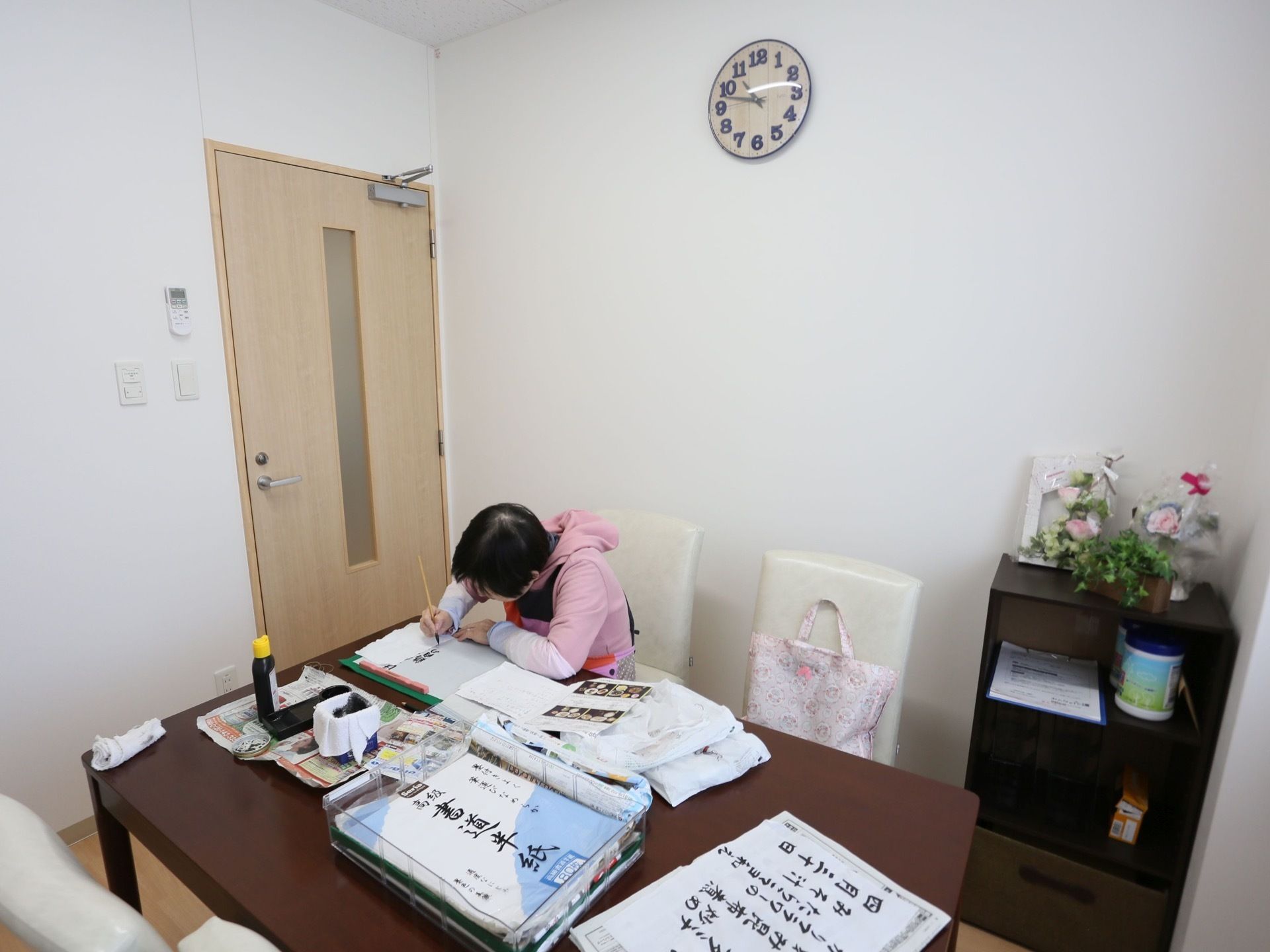 白を基調としたシンプルな部屋で女性が書道をしている。壁に時計、棚に生花が飾られている。机や棚の角が丸く、安全に配慮されている。