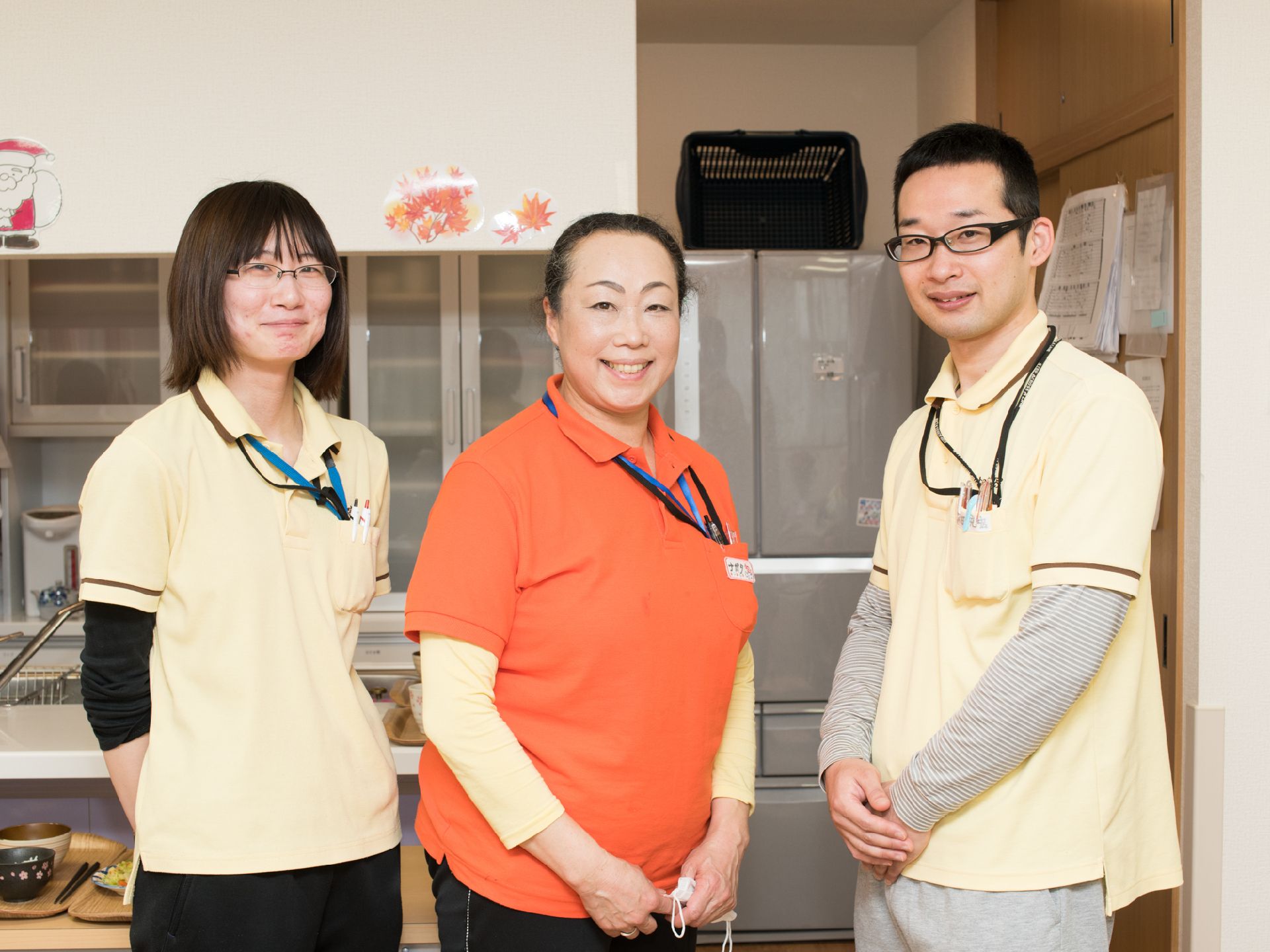 大きな冷蔵庫や食器棚の前に並ぶ女性２人、男性１人のスタッフ
