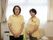 窓際に2人の職員が横並びに立っている。その後ろにはソファが置かれており、その右側には空気清浄機がある。