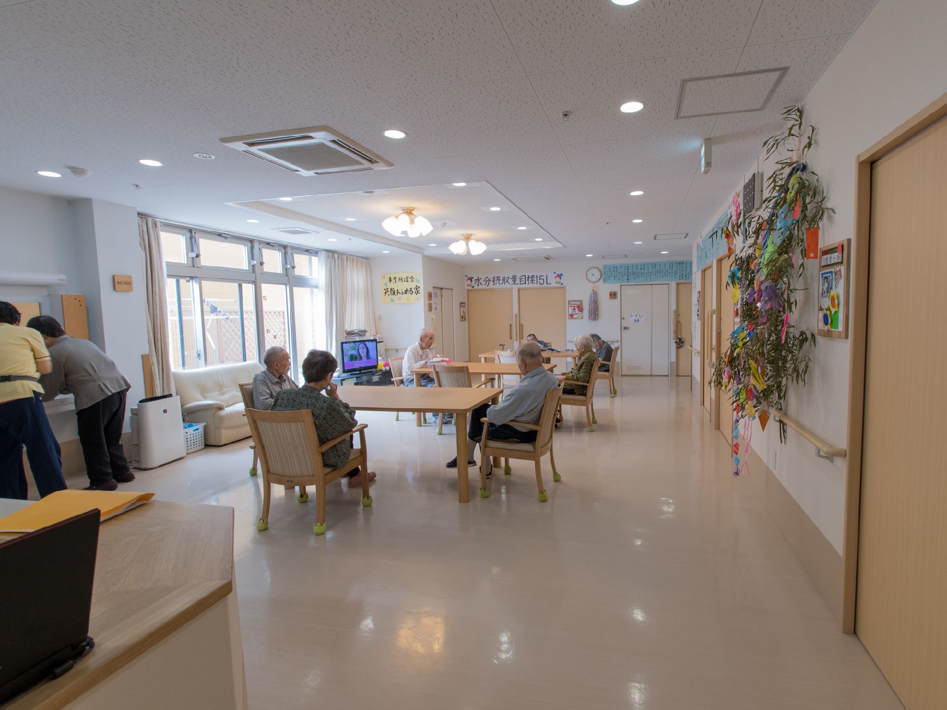 複数のテーブルと椅子が置かれており、椅子には入居者が腰を掛けている。左奥には窓があり、その手前にはテレビが置かれている。