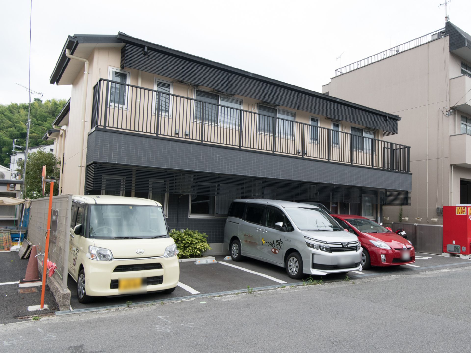 正面に建物が建てられており、その左手前には植え込みがある。周辺は住宅街になっている。奥には山がある。