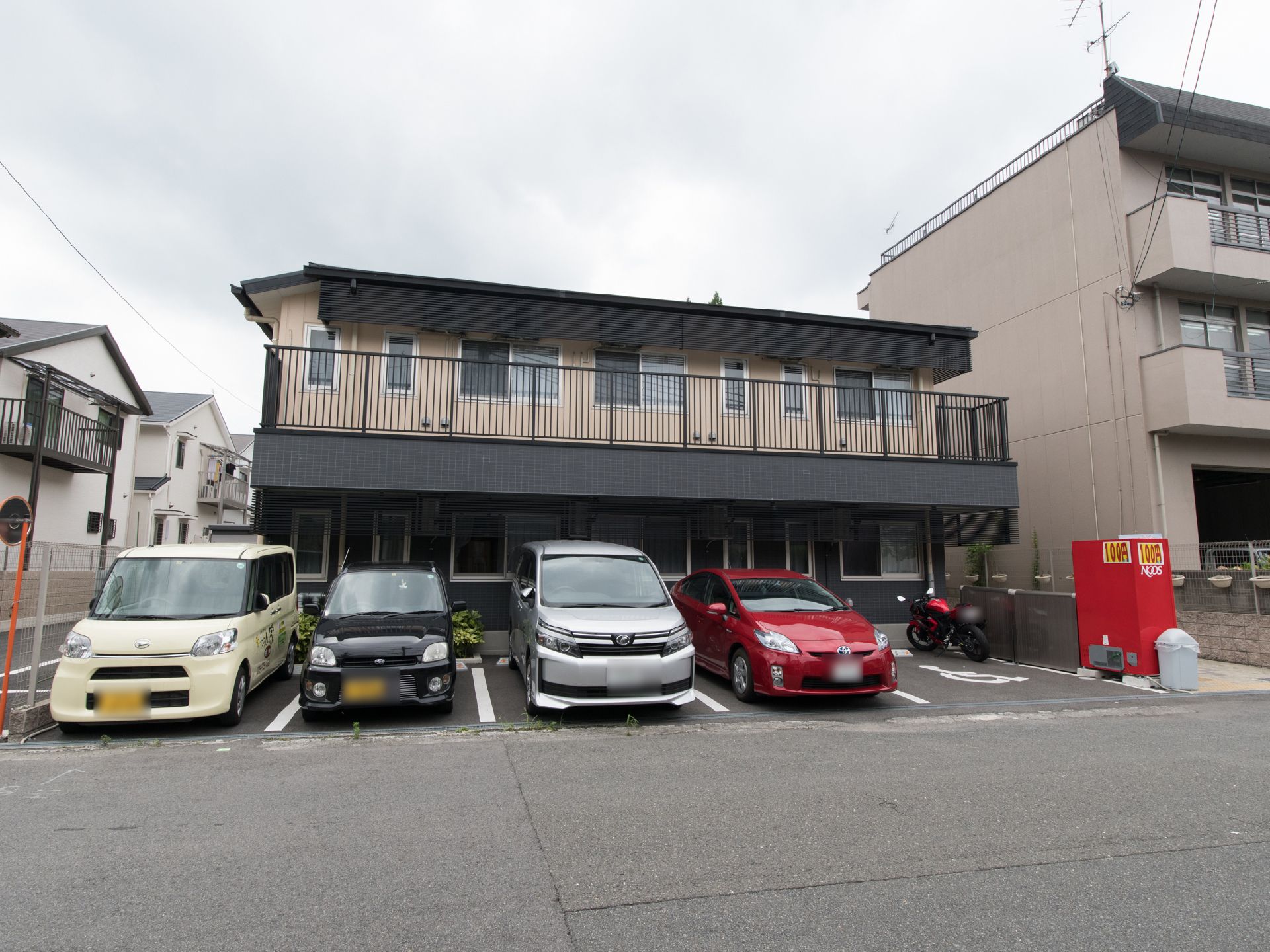 建物が正面に建てられており、その手前には道路がある。1階と2階には複数の窓が付いている。右側には自動販売機がある。