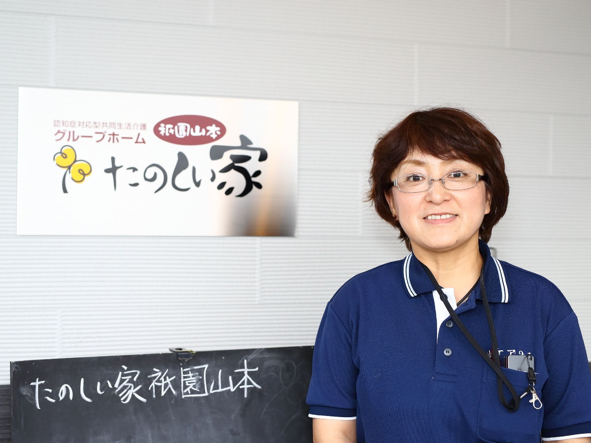 施設の外壁に設置された看板の横で微笑む女性スタッフ