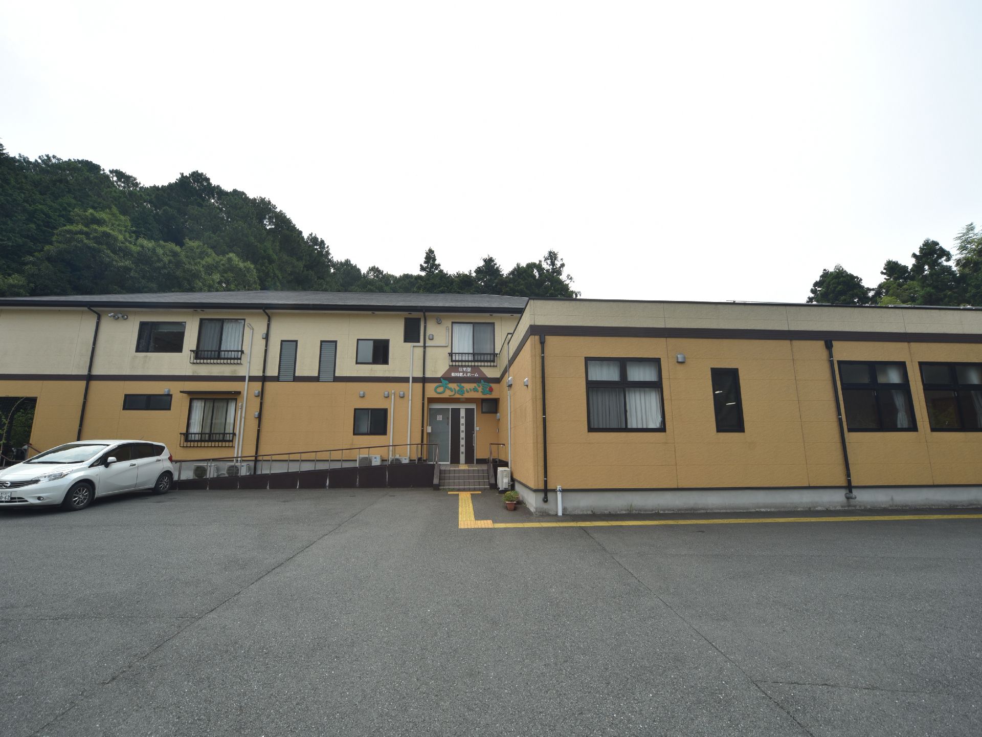 施設の外観は淡いツートンカラーで手前側は平屋で奥が2階建になっている。また、山あいに面した立地である。