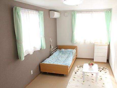 窓が２か所あり、それぞれレースカーテンと緑のカーテンが付けられている。介護ベッドが隅にあり、小さなテーブルと衣装ケースが用意されている。