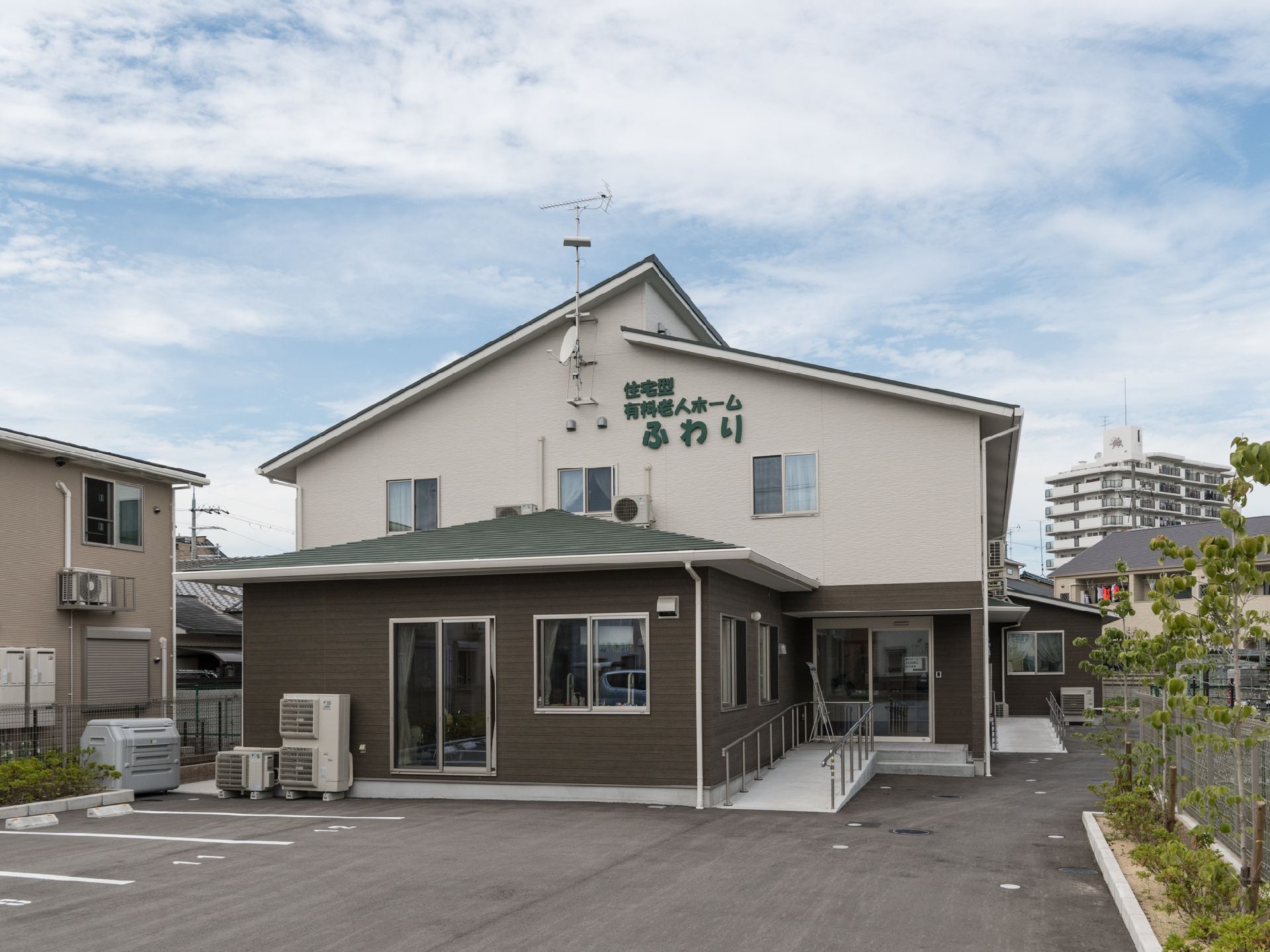 茶色と白色の外壁に、施設名ロゴと屋根が緑色の施設外観は周辺環境にとても馴染んでいる。駐車場から入り口まで広々した空間。