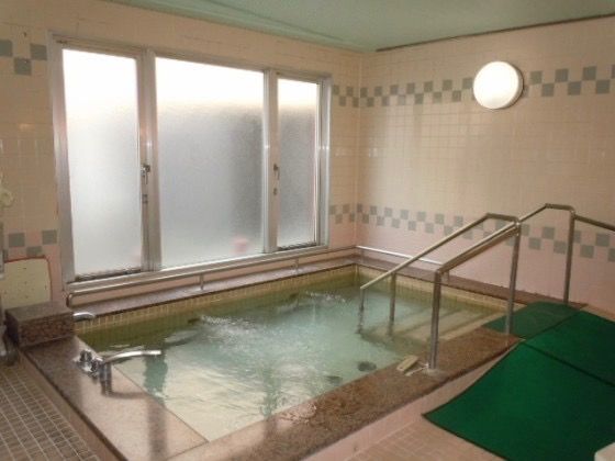外の光が大きなすりガラスの窓から差し込む明るい大浴場である。転倒防止のマットと浴槽周りに手すりを配置している。