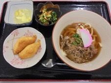 かまぼこ入りの肉うどん、いなり寿司２個、野菜とデザートの小鉢２種類のメニューになっている。手元にはスプーンが置かれている。