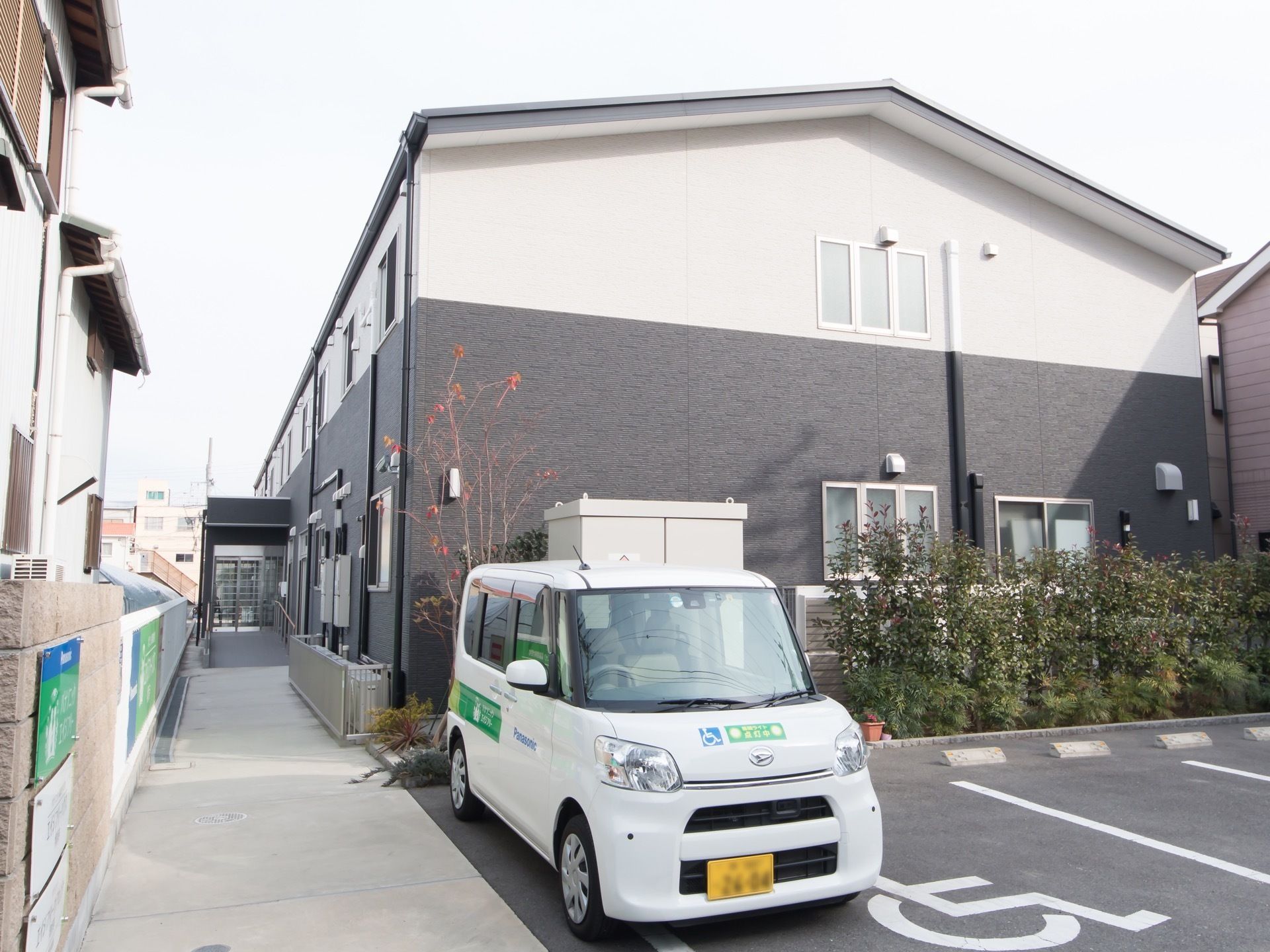 ツートンカラーの外壁の２階建ての建物である。駐車スペースには１台の車が停まっており、軽自動車である。ドアには施設名が書かれている。