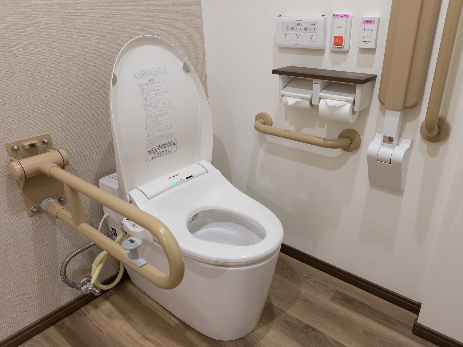 白い壁のトイレである。壁には、薄茶色の手すりが設置されているほか、ペーパーホルダーと緊急呼び出しブザーもある。