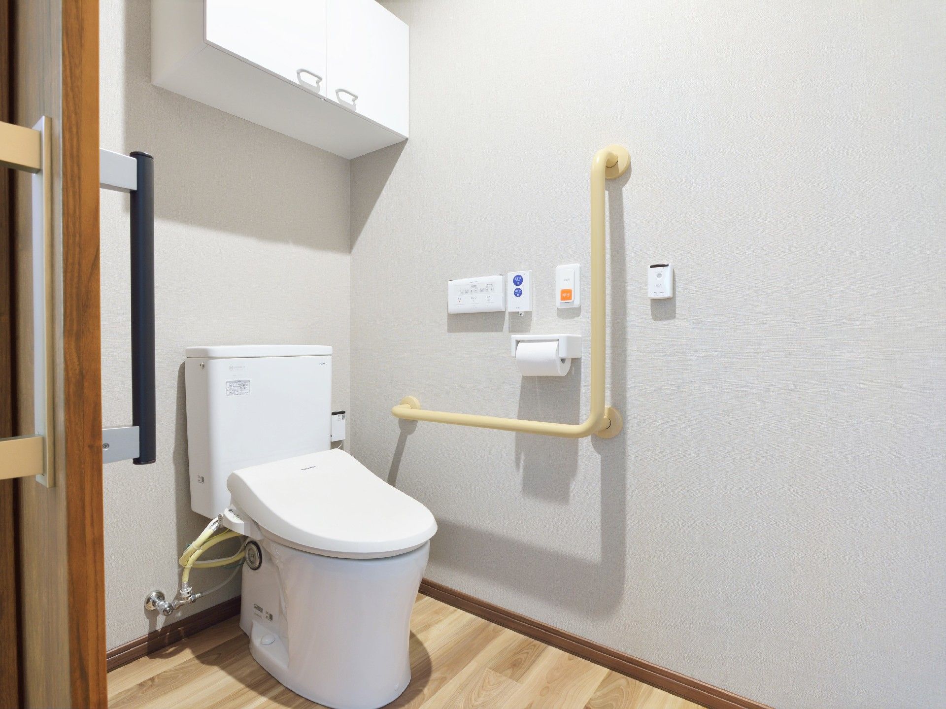 収納棚付きのトイレはフローリングでエル字型の手すりや呼び出しボタンもある。