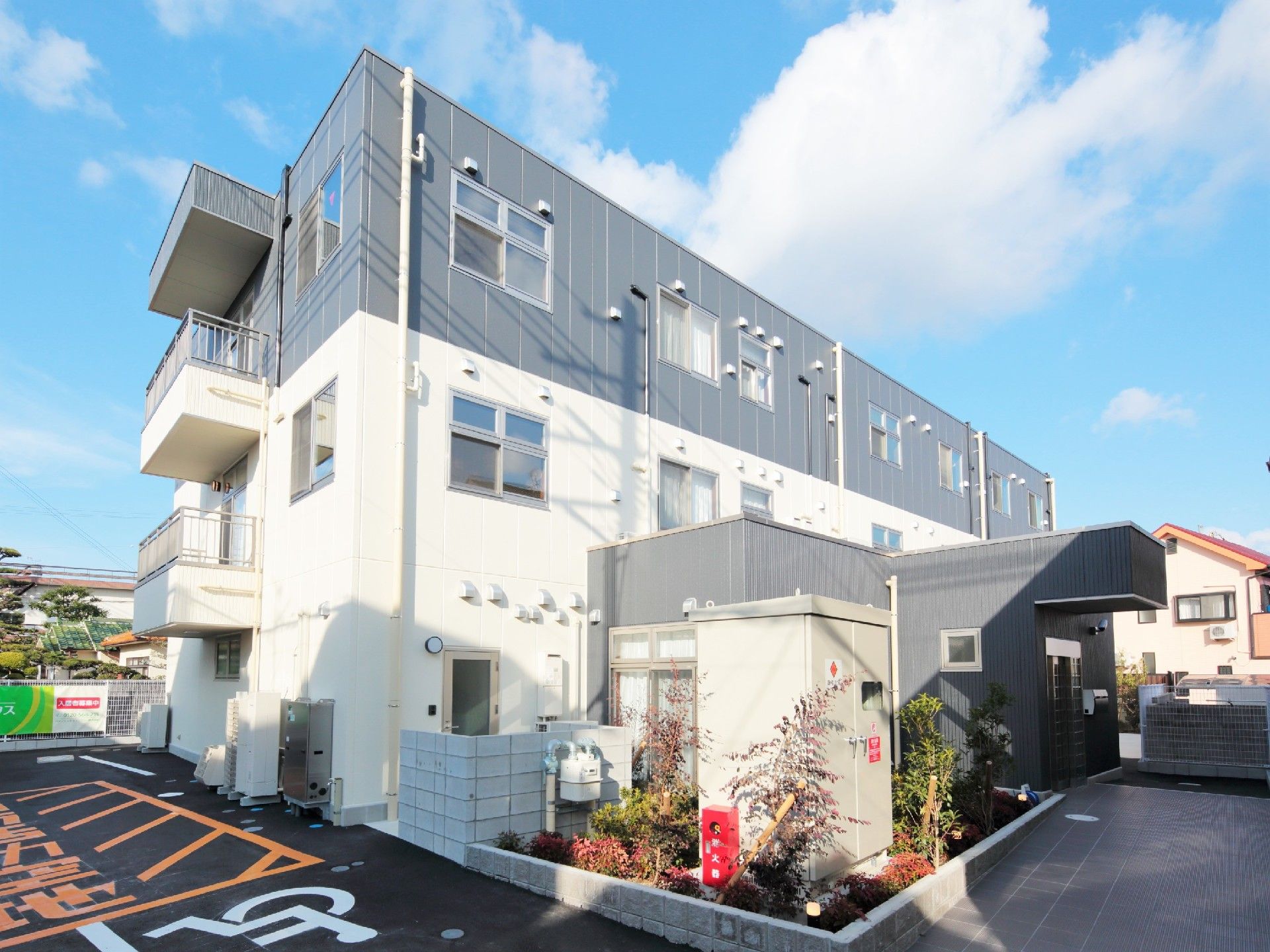 3階建ての建物の1階2階部分は白、3階部分はグレーで、玄関周りには気が植えられている。