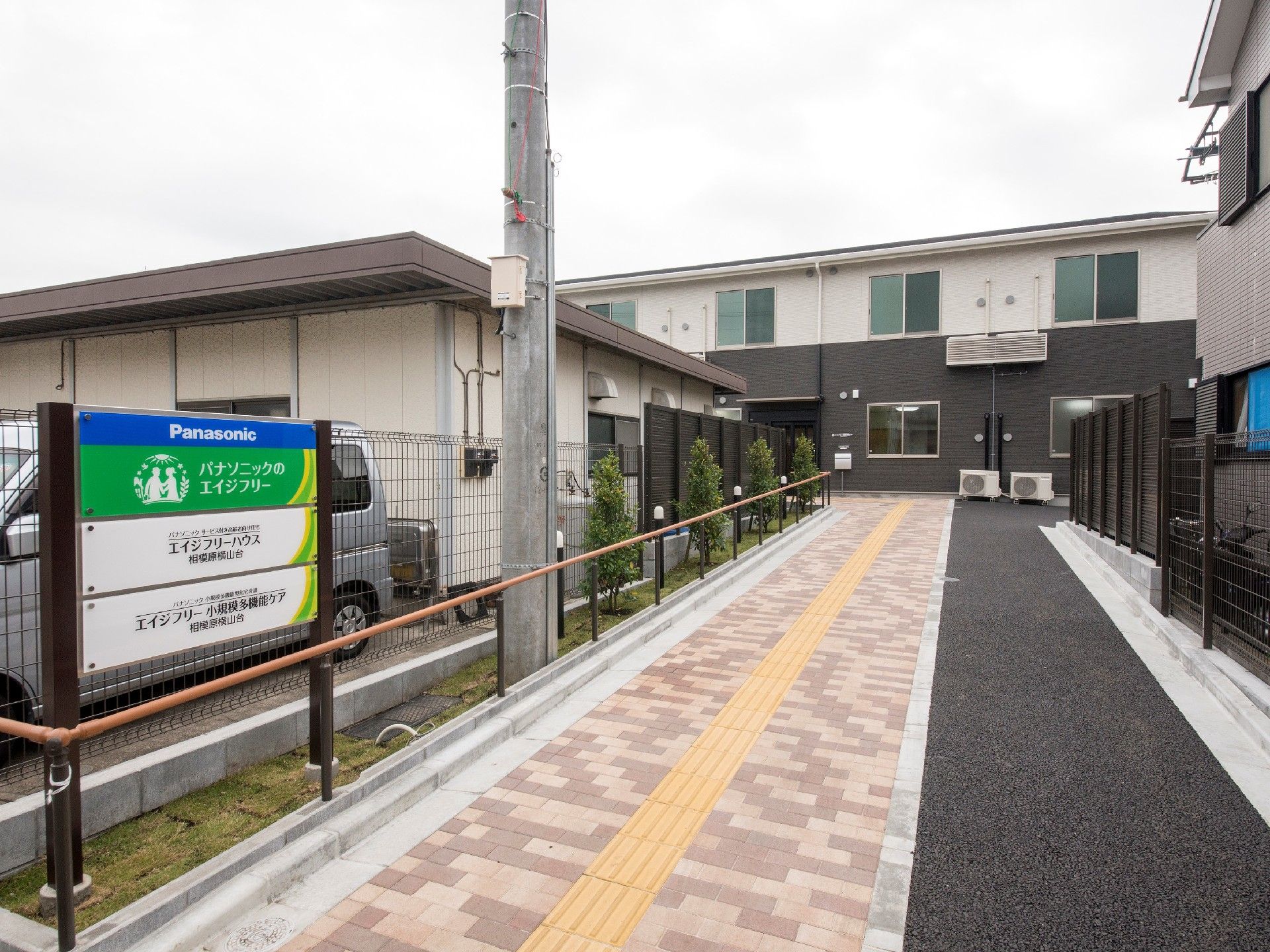 2階建ての建物に続くアプローチは長く、点字ブロックも敷かれている。右側には看板が立っている。
