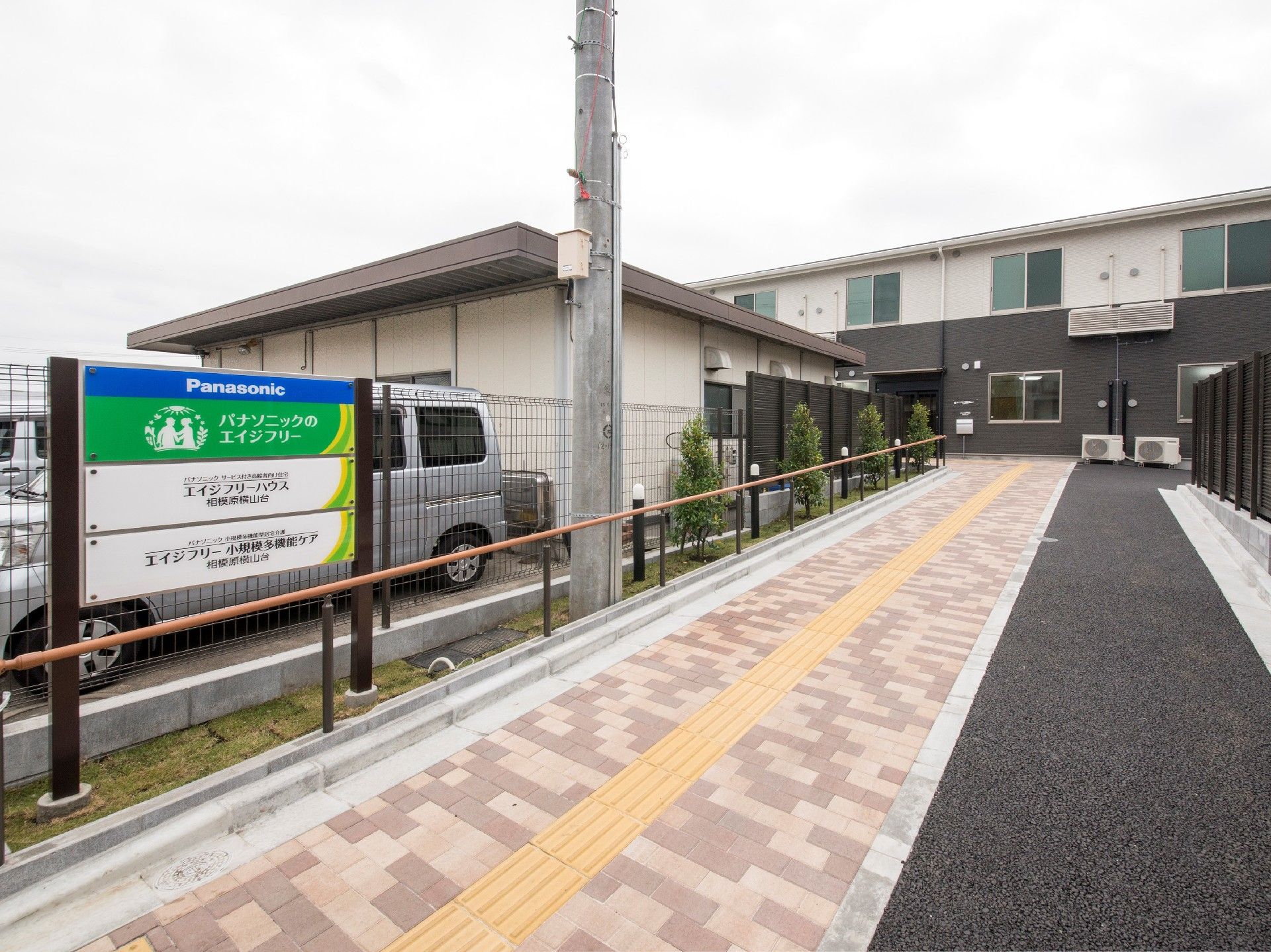 点字ブロックが敷かれたレンガ調のブロックのアプローチが2階建ての建物に続いている。
