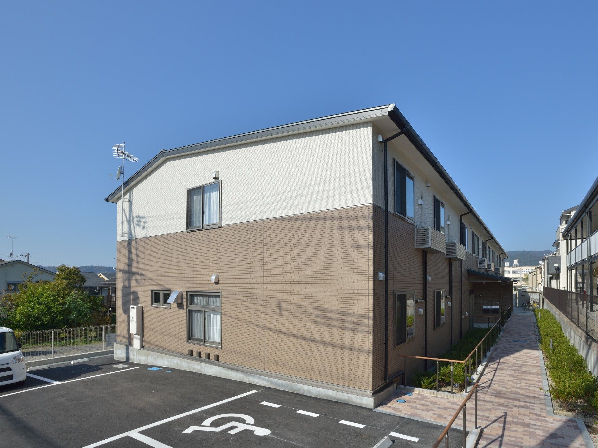 白とブラウンの2階建て他の建物。玄関には手すり付きのスロープがあり、広い駐輪場もある。