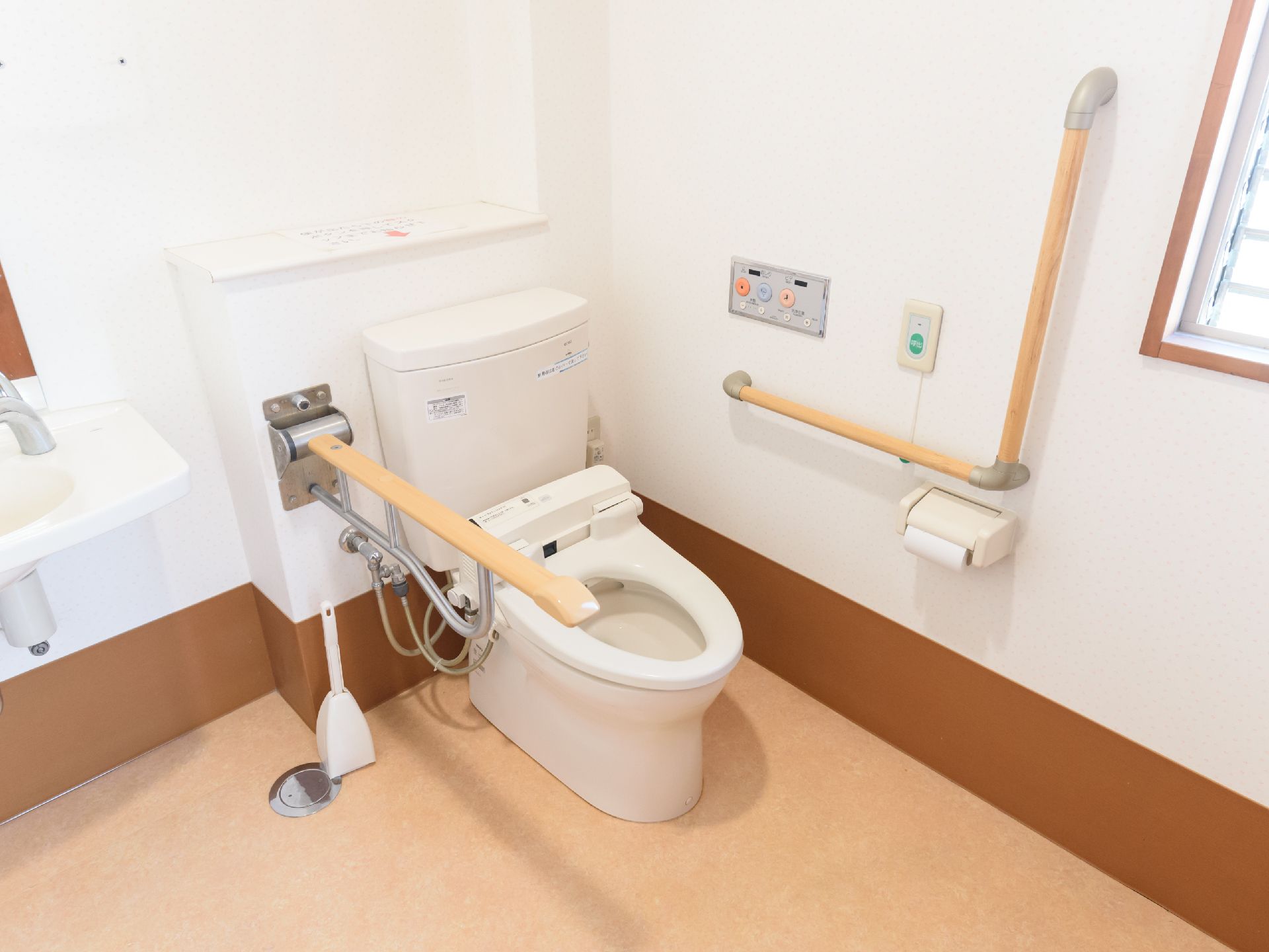 白い壁の清潔感のある個室トイレで、壁にＬ字型の手すりが設置されている。手すりの近くにはナースコールが取りつけられている。