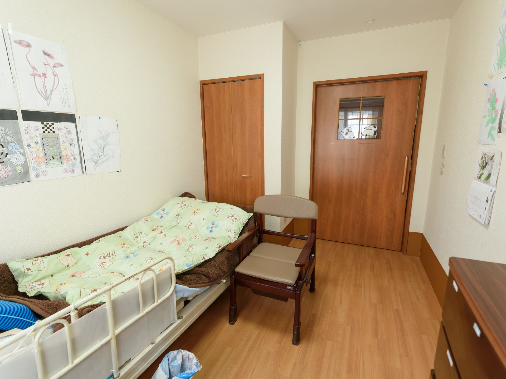 壁側にベッドが１台設置されており、その脇にポータブルトイレが置かれている。壁にはカレンダーや塗り絵などが飾られている。