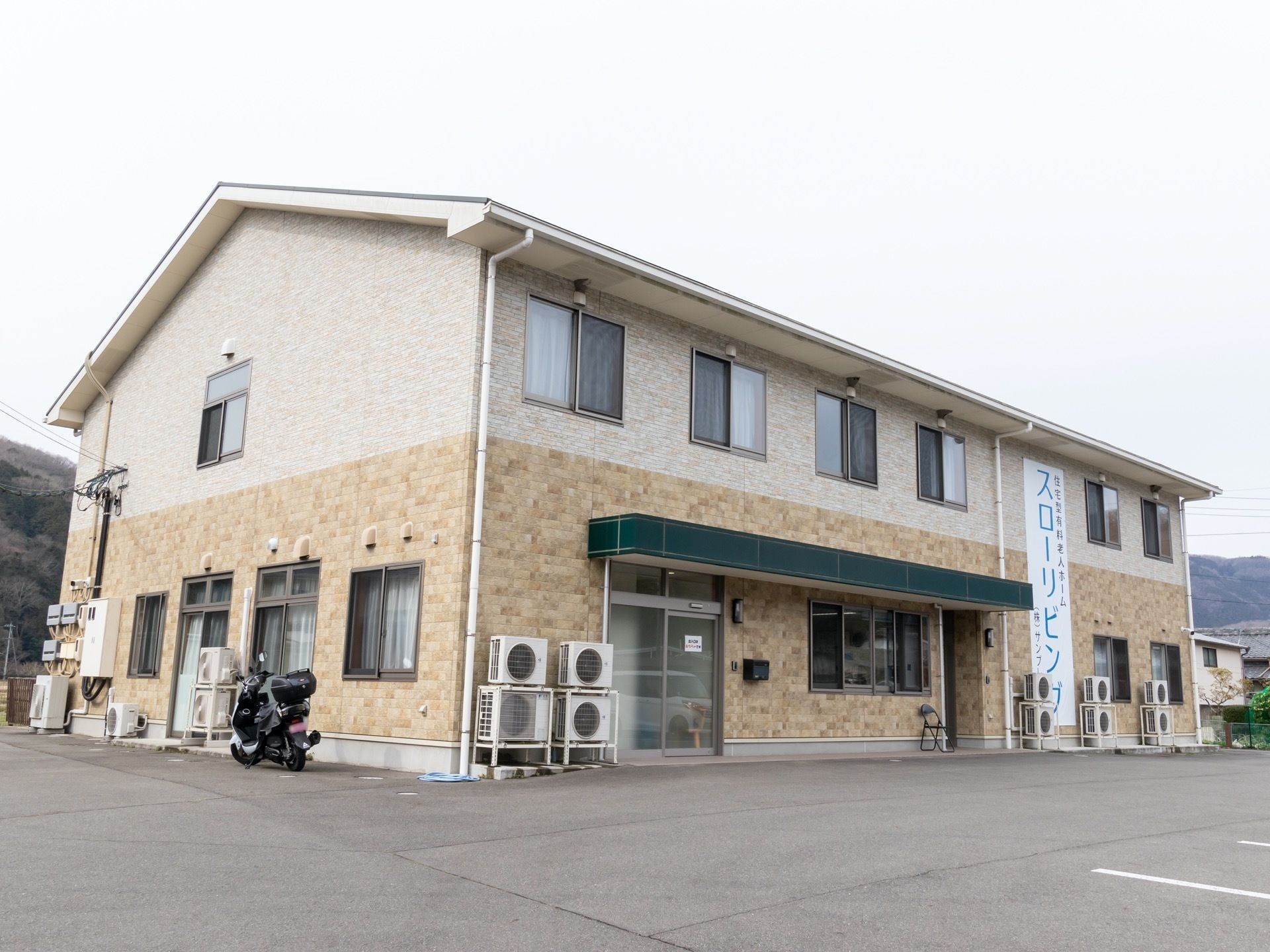 広い駐車場が完備されている施設で、外壁沿いにはアコンの室外機が複数台設置されている。周辺には緑豊かな山々が見える。